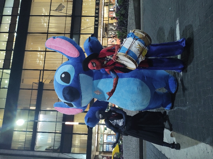 The image captures three costumed figures posing at night on a paved urban surface in front of the Teatro San Martín in Buenos Aires, Argentina. Centrally positioned is a large, inflatable blue figure resembling Disney's Stitch, featuring prominent pink inner ears and a light blue abdominal patch with a red emblem. To its left, an individual is dressed in a black full-body costume with white markings, characteristic of Venom, a black mask, and a red scarf, paired with a long black skirt or flowing trousers. This person's left arm is extended outward with an open palm. To the right of the Stitch figure, another individual wears a red and blue costume reminiscent of Spider-Man, accentuated by a black cowboy hat, a red scarf or cape, and wide, lustrous blue pants. This figure holds a large drum decorated with the Argentinian flag design, including the sun emblem, and performs a hand gesture with their right hand. The background consists of a multi-story modern building with numerous illuminated glass windows, behind which white curtains are visible, and partial signage reading "TEATRO S" is discernible. A bright, vertical streetlamp is situated behind the central Stitch character. The ground is composed of dark, interlocking pavers, exhibiting some reflective areas. Distant street activity, including blurred vehicles and individuals, is visible behind a low-lying planter containing green foliage.
