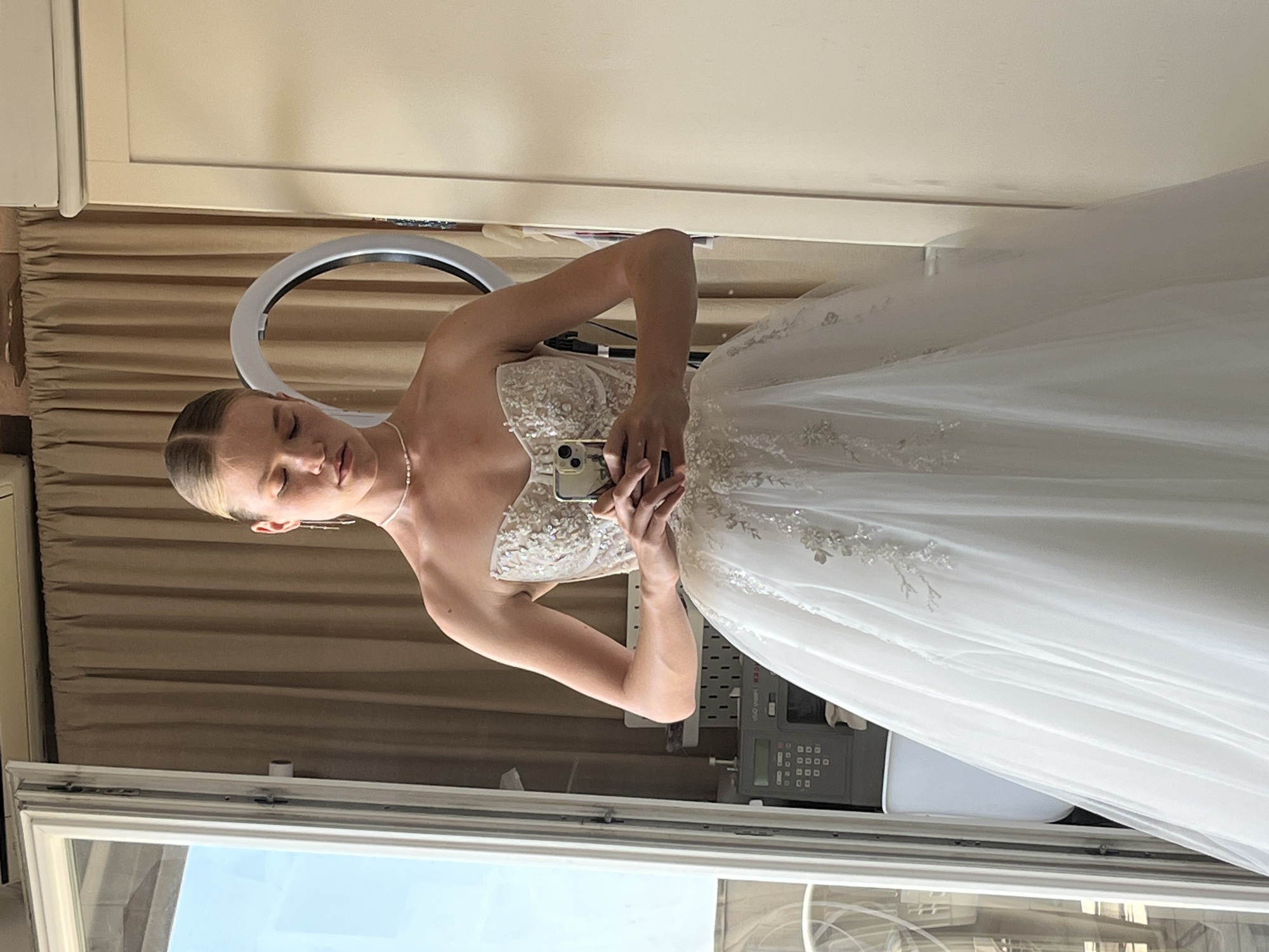 A young woman stands in a white strapless wedding dress, holding a smartphone to take a selfie. Her eyes are closed and her head is tilted down. She is wearing a delicate pearl necklace. The background is a room with beige curtains, a large mirror reflecting the scene, and a white cabinet. To the left, a window shows a glimpse of an outdoor space with a view of a city, possibly Budva, Montenegro. A sewing machine is visible to the left of the woman. A ring light is positioned behind her to the right. The lighting suggests an indoor setting, potentially a fitting room or a hotel room.