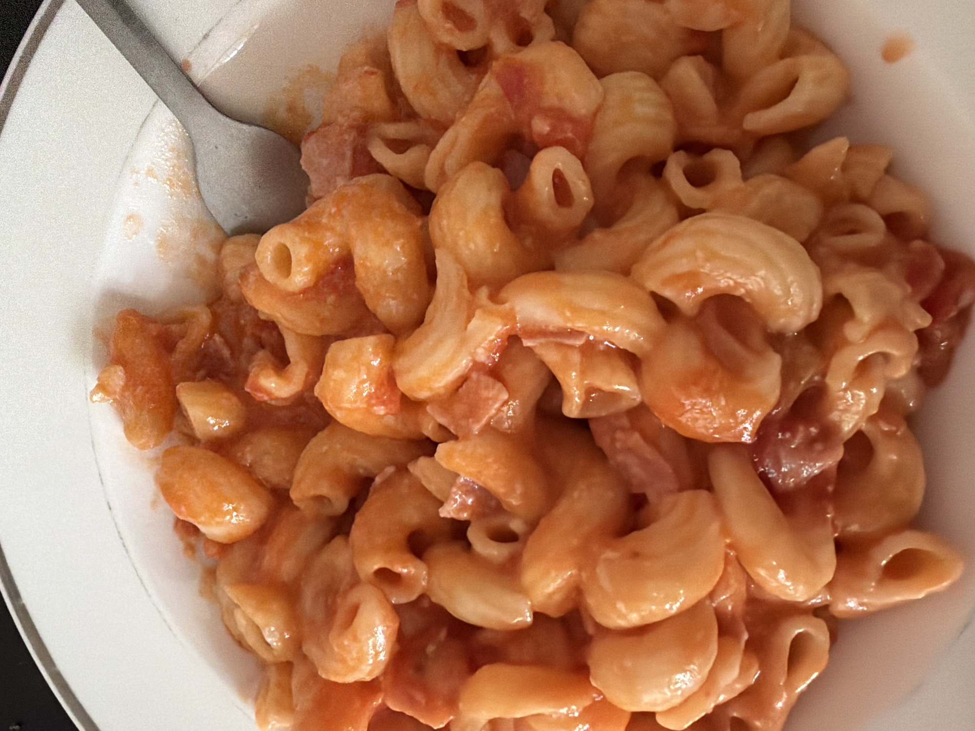 The image depicts a close-up of a bowl of pasta, specifically elbow macaroni, coated in a reddish-orange sauce. A metal spoon is partially visible on the left side of the frame, positioned within the bowl. The pasta is piled high in the white bowl, with the focus primarily on the texture and color of the dish. The overall lighting is bright, accentuating the sheen of the sauce. There is no indication of location in Pentre, United Kingdom, or any people present.