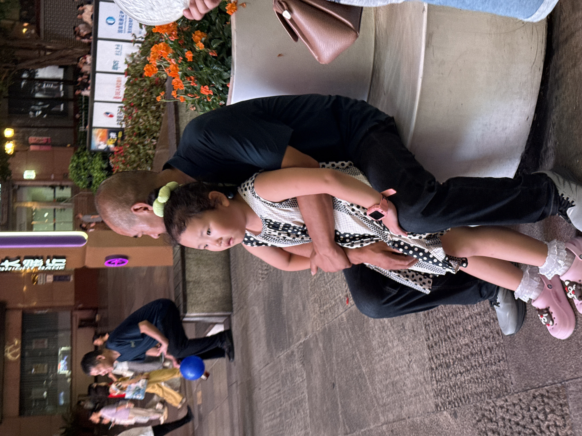 The image shows a father and daughter sitting on a paved area at what appears to be dusk or night. The father is wearing a black t-shirt and black pants, with his arm around his daughter. The daughter is wearing a black and white polka dot dress and pink shoes with bear decorations and white lace socks. She is looking towards the camera. In the background, there are illuminated signs with Chinese characters and some orange flowers. Further in the background, other people can be seen walking. A blue ball is visible on the ground near some people in the background. The scene suggests an outdoor setting, possibly a street or public square, in an urban environment in Yuzhong District, China. The overall mood appears calm and familial.