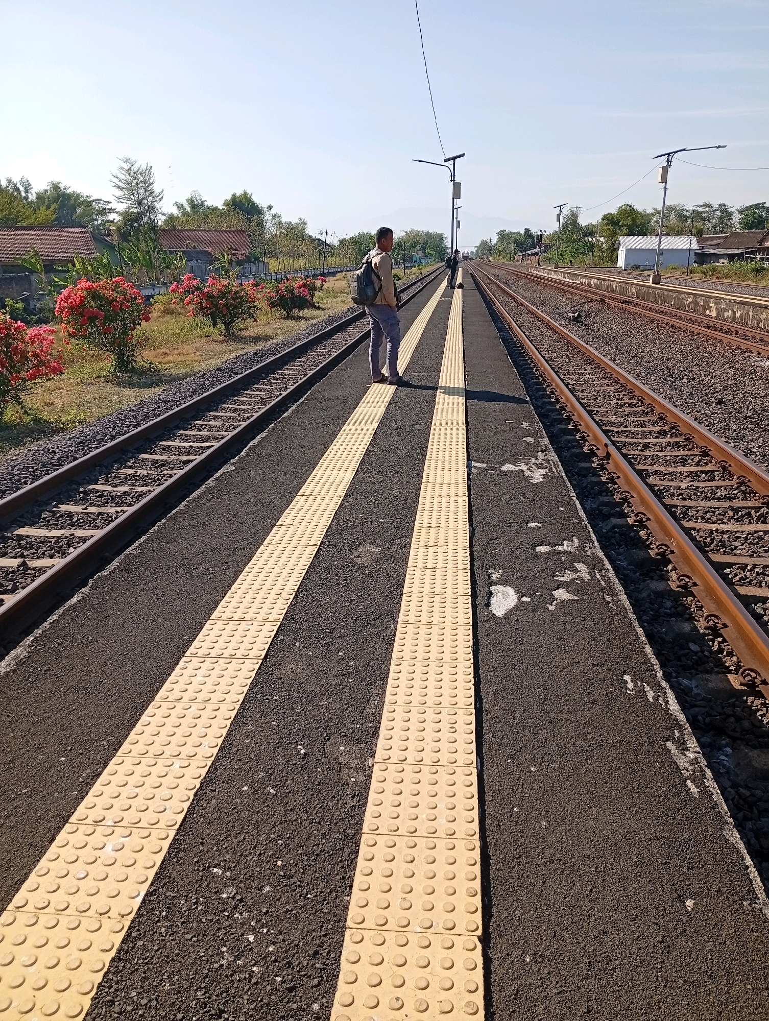The image shows a man standing on a train platform in Tebon, Indonesia. The platform has two yellow tactile paving strips running parallel to the train tracks. The man is wearing a light-colored shirt, dark pants, and a backpack. He is looking down the train tracks. The train tracks are made of gravel and metal. There are trees and houses in the background. The sky is clear and blue.