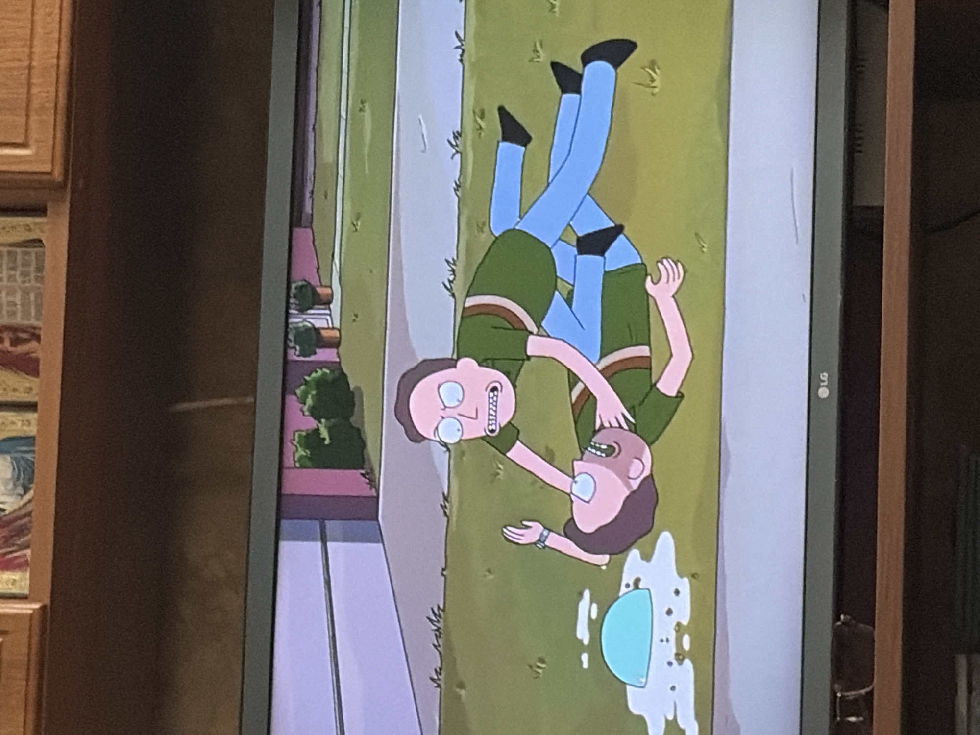 The image shows a cartoon depiction of two figures in a yard. The figure on top, with light brown hair and a pinkish complexion, is wearing a green shirt and blue pants. This figure is straddling the figure below, with its hands around the neck of the other. The figure below, with dark brown hair and a paler complexion, is lying on its back and wearing a similar green shirt and blue pants. Its eyes are wide and its mouth is open in a distressed expression, with one hand reaching up towards the figure above. A light blue, dome-shaped object is on the ground to the left of the figures, surrounded by a white, liquid-like substance. The figures are on a green lawn with sparse blades of grass. In the background, there is a light grey driveway and a house with pink siding and white trim. Green bushes are planted in front of the house, and two potted plants are visible. The scene appears to be during daylight.
