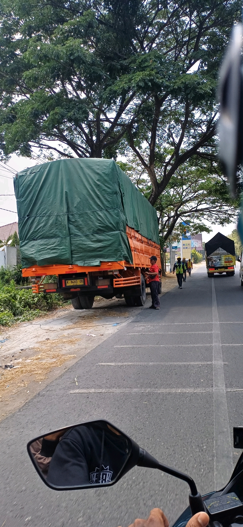 A truck covered in green tarp is parked on the side of a road in Kecamatan Pati, Indonesia, with a man in a red shirt standing near its rear. The truck, an orange flatbed with dual rear wheels, has a license plate reading "K-8465 OB." In the background, other vehicles, including a yellow truck with a black tarp, are driving on the asphalt road. Tall trees with dense green foliage line the right side of the road, and a building with a white fence is visible on the left. The image is taken from the perspective of someone riding a motorcycle, as evidenced by a mirror reflecting the rider's black hoodie and a hand gripping a handlebar. The road surface is marked with white lines, and the shoulder is dusty with scattered debris.