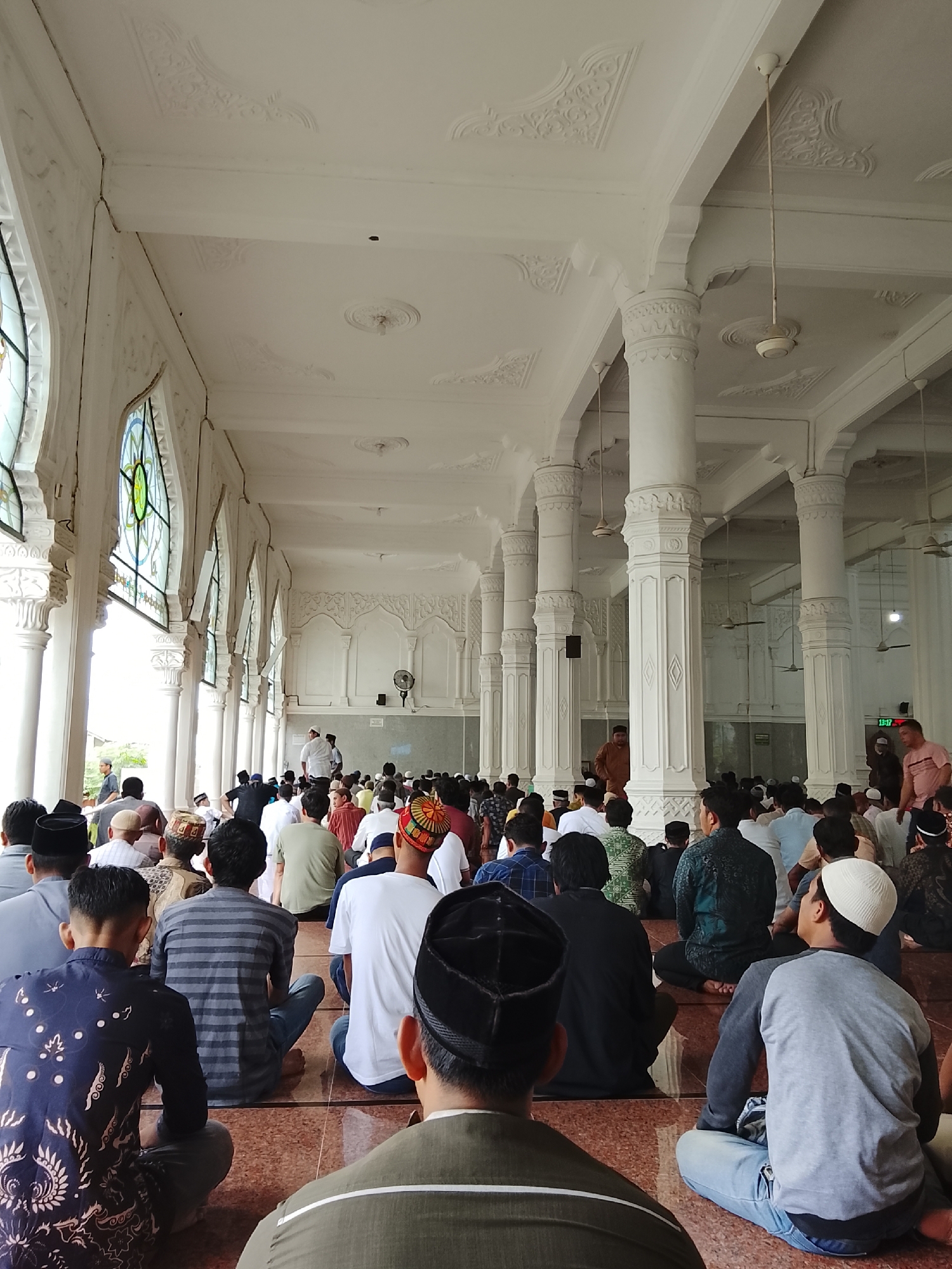 The image depicts a congregation of Muslim men inside a mosque in Lhokseumawe, Indonesia, during prayer. The men are seated on a reddish-brown tiled floor, facing a white wall. They are dressed in various traditional Islamic attire, including peci hats, sarongs, and shirts. The mosque architecture features white columns, ornate ceiling decorations, and arched windows with stained glass. Some ceiling fans are visible. In the background, a few individuals are standing, possibly preparing for prayer or interacting with others. The overall atmosphere suggests a religious gathering.