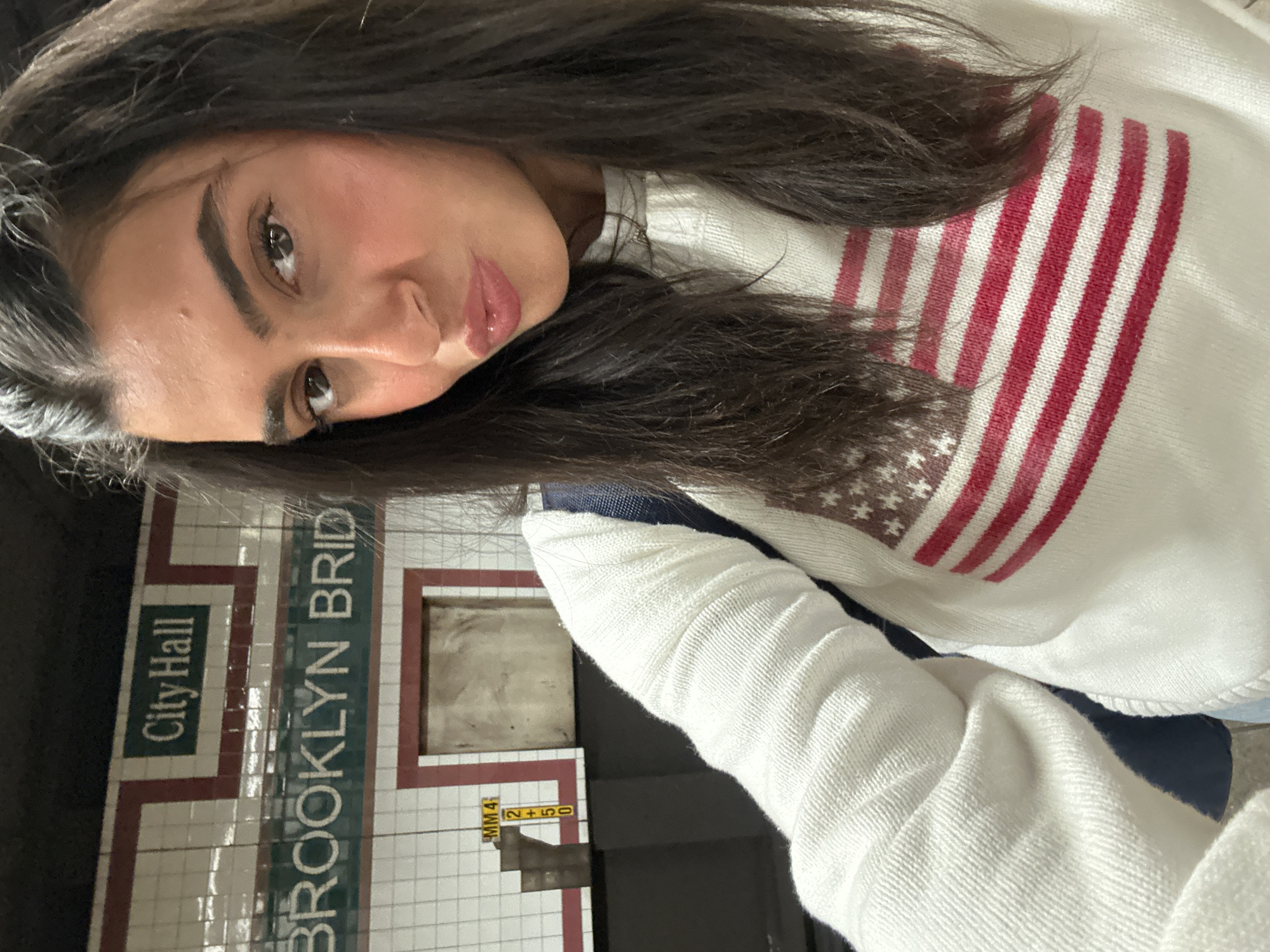 The image depicts a woman with long, dark hair taking a selfie. She is wearing a white sweater with a red and white striped pattern resembling the American flag. The background shows a subway station wall with white tiles and dark green lettering that reads "City Hall" and "BROOKLYN BRIDGE." The station appears to be in the City Hall-Brooklyn Bridge area of New York City. There are also some yellow signs and details that suggest it is a subway platform setting.