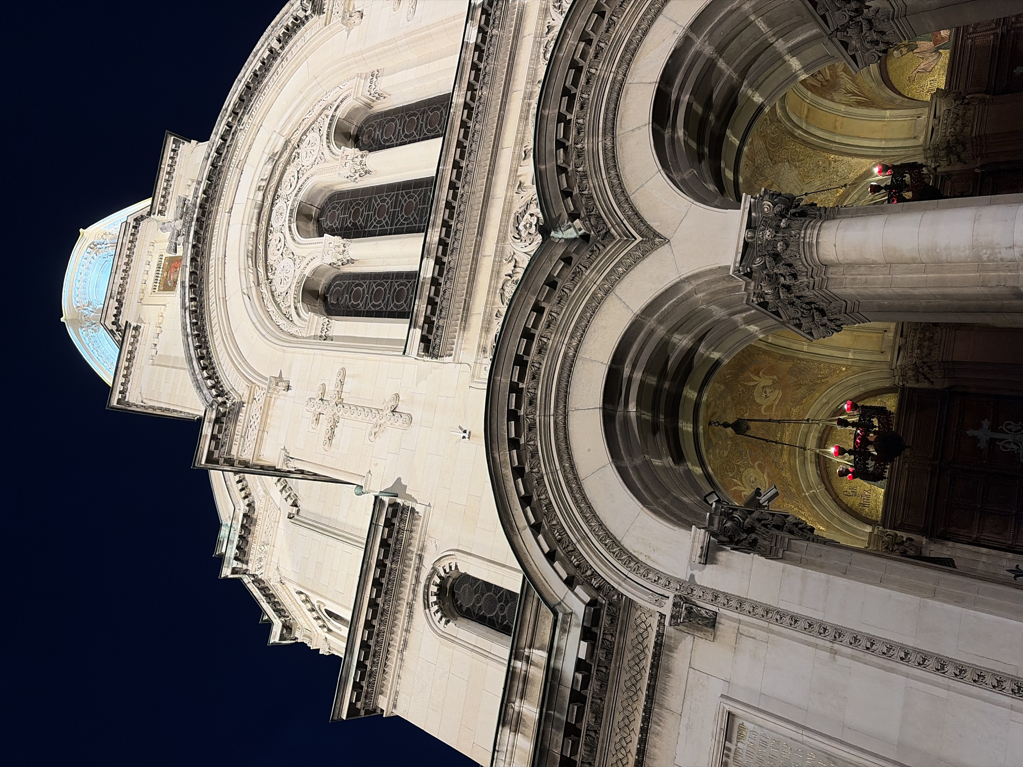 The image showcases a section of a grand building, likely a church or cathedral, at night. The structure is predominantly constructed of light-colored stone, adorned with intricate carvings, arches, and windows featuring complex geometric patterns. A light blue dome is visible on the left. Inside the archways, gold mosaic ceilings and elaborate chandeliers with red lights can be seen. The background is a dark night sky. The perspective is looking upwards at an angle, emphasizing the building's height and architectural details. The location is in the city of Unknown, Unknown.