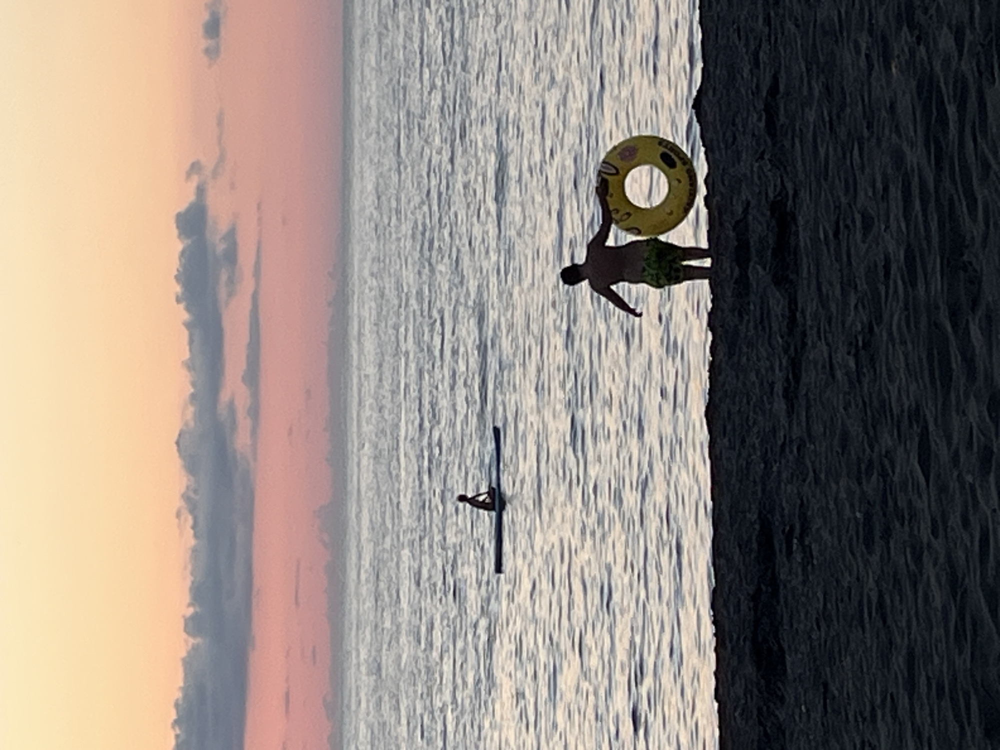 At dusk in Ureki, Georgia, a serene beach scene unfolds. The sky transitions from a pale orange to a soft pink, with scattered grey clouds adding texture. The sea, a calm expanse of rippling water, reflects the soft light. In the distance, a lone figure is seated on a paddleboard, seemingly enjoying the tranquility. Closer to the shore, a person stands on the sandy beach, holding a yellow inflatable ring. The foreground is dominated by dark, granular sand, suggesting the typical composition of Ureki's magnetic sand beaches. The overall atmosphere is one of peaceful evening relaxation by the Black Sea.