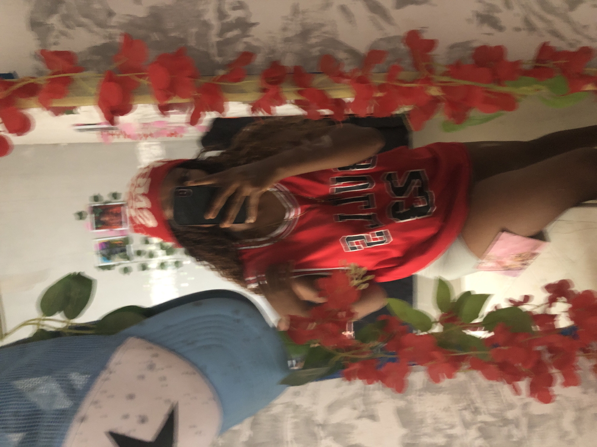 A person is reflected in a mirror, holding a smartphone to take a selfie. The person is wearing a red jersey with the number "23" on it. There's a blue baseball cap with a black star on the lower left of the mirror's reflection. Red artificial flowers are draped around the edges of the mirror. The background shows a neutral-colored wall with a pattern that resembles fabric. The scene does not provide enough evidence to determine the location in Kashere, Nigeria.