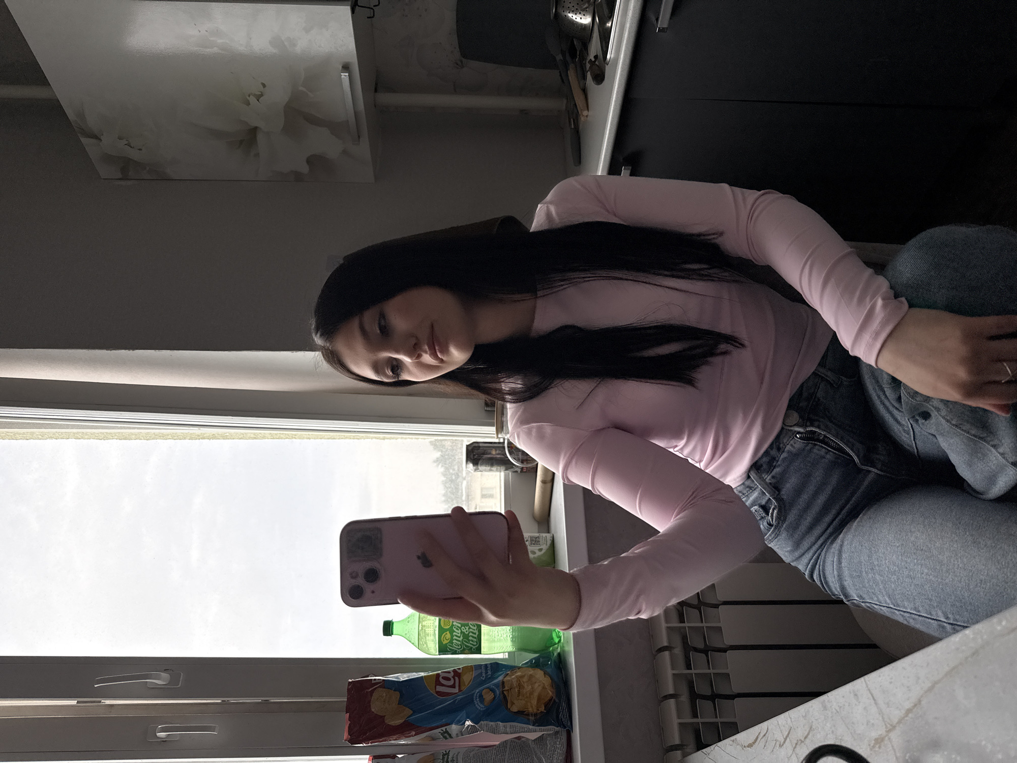 An interior scene depicts a young adult female seated in what appears to be a kitchen or dining area in Chaikovsky, Russia. The subject, possessing long dark hair, wears a light pink long-sleeved top and blue jeans. She is positioned facing slightly away from the camera, holding a pink smartphone vertically in her right hand, seemingly interacting with its screen. A silver ring is visible on the ring finger of her left hand, which rests on her lap.
To the left of the subject, on a windowsill, are a bag of Lay's potato chips (Sour Cream & Herbs flavor, indicated by Cyrillic text), and a green plastic bottle of "Lemon & Lime" soda. To the right of the subject, also on the windowsill, is a black can and a cylindrical wooden object. A white multi-pane window dominates the background, offering a view of an overcast sky and distant structures consistent with an urban or suburban environment. A white radiator is visible below the window.
To the right of the subject, dark gray/black kitchen cabinets with silver handles are visible, alongside a white wall-mounted cabinet featuring a faint floral design. A countertop below the cabinets holds various kitchen items, including a metal colander, a dark cutting board, and other utensils. A portion of a sink area is also discernible on the far right. The room is illuminated by natural light entering through the large window.