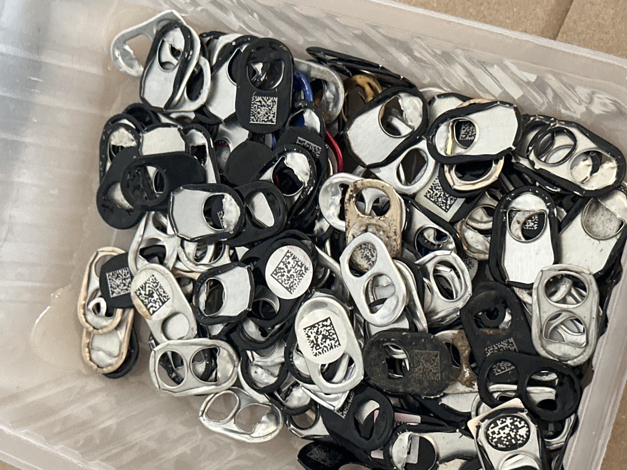 The image shows a close-up, overhead view of a clear plastic container filled with a large quantity of pull tabs from beverage cans. The pull tabs are predominantly silver and black, with some appearing worn or tarnished. Several of the tabs feature small, printed QR codes. The container is made of clear plastic, and its sides are visible along the edges of the image. The background appears to be a textured, light brown surface, possibly cardboard. There are no people or discernible actions taking place within the frame. The scene depicts a collection of discarded items. The location in the city of Nevinnomyssk, Russia is not ascertainable from the visual information provided in the image.