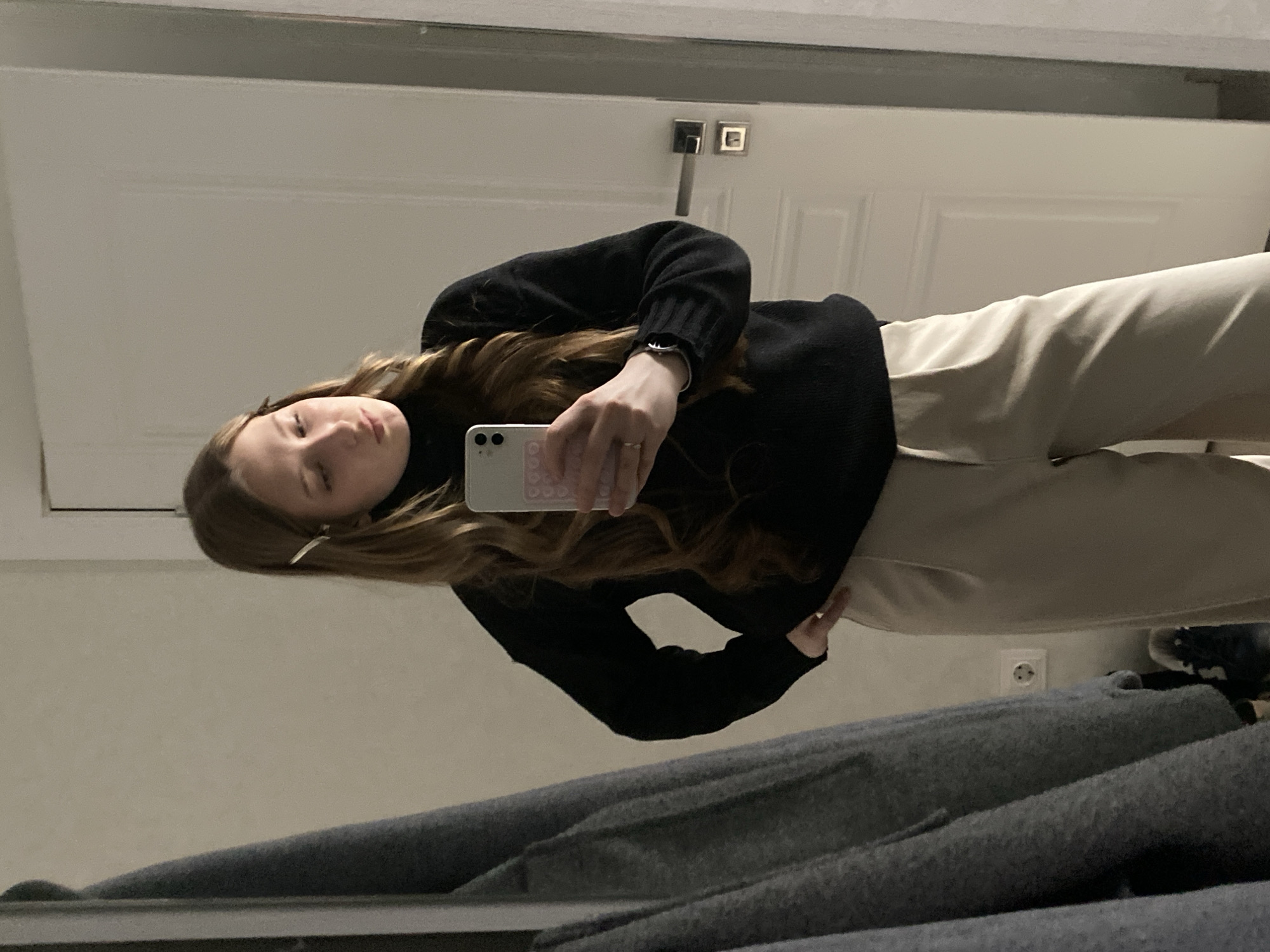 A photograph taken in Kazan, Russia, shows a woman in a black turtleneck sweater and tan pants holding a smartphone with a pink and white case, presumably taking a selfie in a mirror. She has long, wavy brown hair with a silver hair clip. Behind her, there is a white door with silver hardware and a white wall. In the foreground, draped over what appears to be the bottom of the mirror frame, are folded gray garments, possibly pants or blankets. A partially visible wall outlet and footwear are present in the lower right corner. The lighting appears to be indoor and somewhat dim.