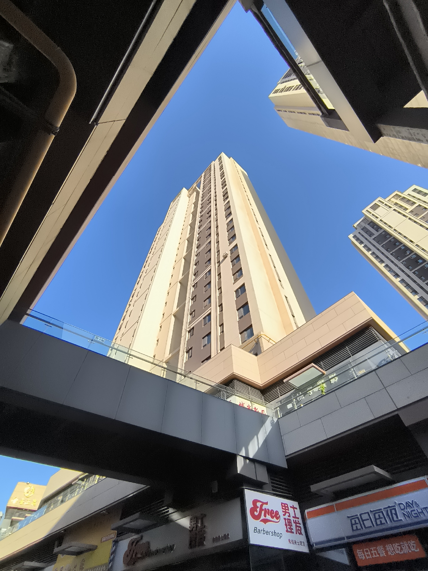 This is an outdoor, low-angle, wide shot of modern architecture under a clear blue sky in Ürümqi, China. The foreground features the undersides of elevated walkways and structures, one with a curved, large diameter pipe. These structures frame a view looking up at two tall, contemporary buildings. The primary building in the center is a light-colored skyscraper with many windows and a repeating vertical pattern of darker sections. To the right, another, slightly shorter building of similar design is visible. Below the elevated walkways, storefronts are visible, including signs for "Free Barbershop" and "每日商务 DAY & NIGHT". The overall impression is of an urban commercial or residential district with multi-level infrastructure. There are no people or animals visible in the frame.