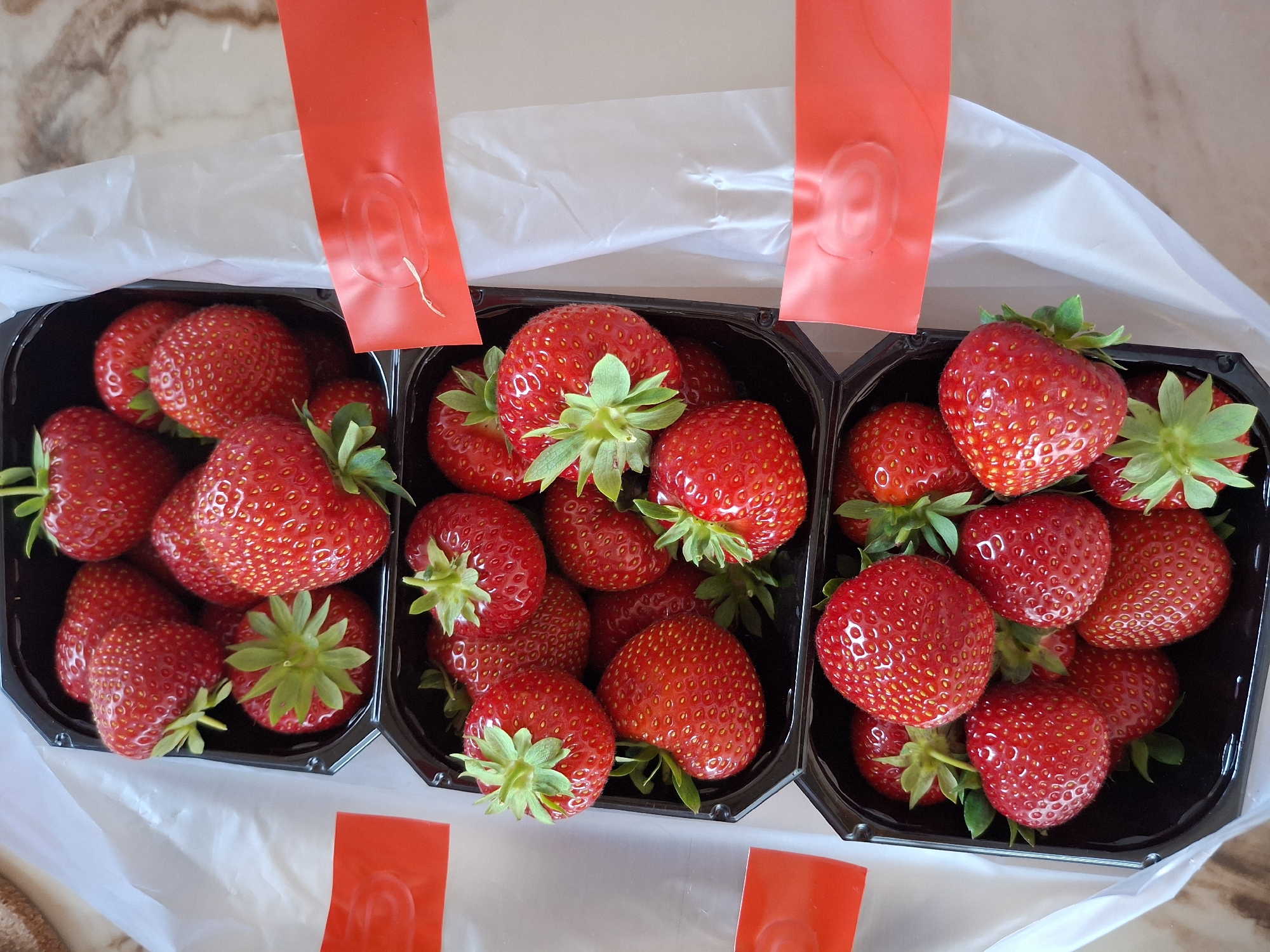 The image shows three black plastic punnets filled with ripe strawberries, arranged side-by-side. Red paper tabs are visible, folded over the edges of the punnets, suggesting they are part of packaging or a display. The strawberries are a vibrant red, with visible seeds and green leafy tops. The punnets are placed on a light-colored surface, possibly a countertop or table, with a marble-like pattern visible in the background. A white plastic bag or liner is also apparent, surrounding the punnets. The lighting suggests it is daytime, with natural light illuminating the scene. The overall impression is one of fresh produce, likely ready for sale or consumption. There is no visible text in the image. The location context provided is Åsgårdstrand, Norway.