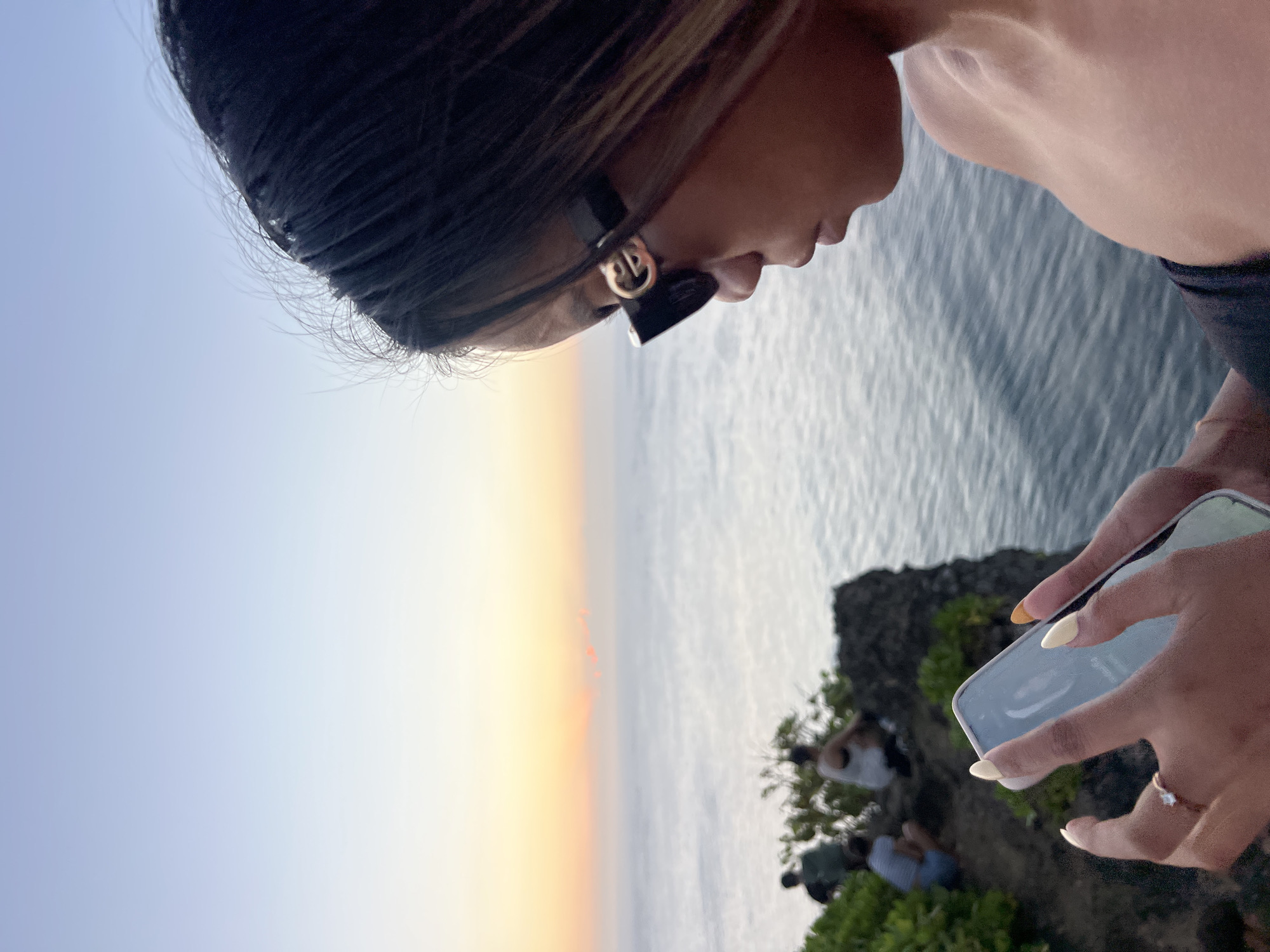 A young woman with dark hair and tan skin, wearing black designer sunglasses and a strapless black top, is holding a smartphone in her hands. She is looking down at the phone, with her face in profile. She is wearing a ring on her left ring finger, and her fingernails are painted yellow and orange. The background shows a calm ocean during sunset, with a soft gradient of orange and blue in the sky. In the middle ground, on a rocky cliff overlooking the ocean, are three other people, appearing to be tourists. They are dressed casually and appear to be looking out at the sunset. The overall setting suggests a scenic overlook in the popular tourist area of Kuta Selatan, Bali, Indonesia, known for its beautiful beaches and sunset views.
