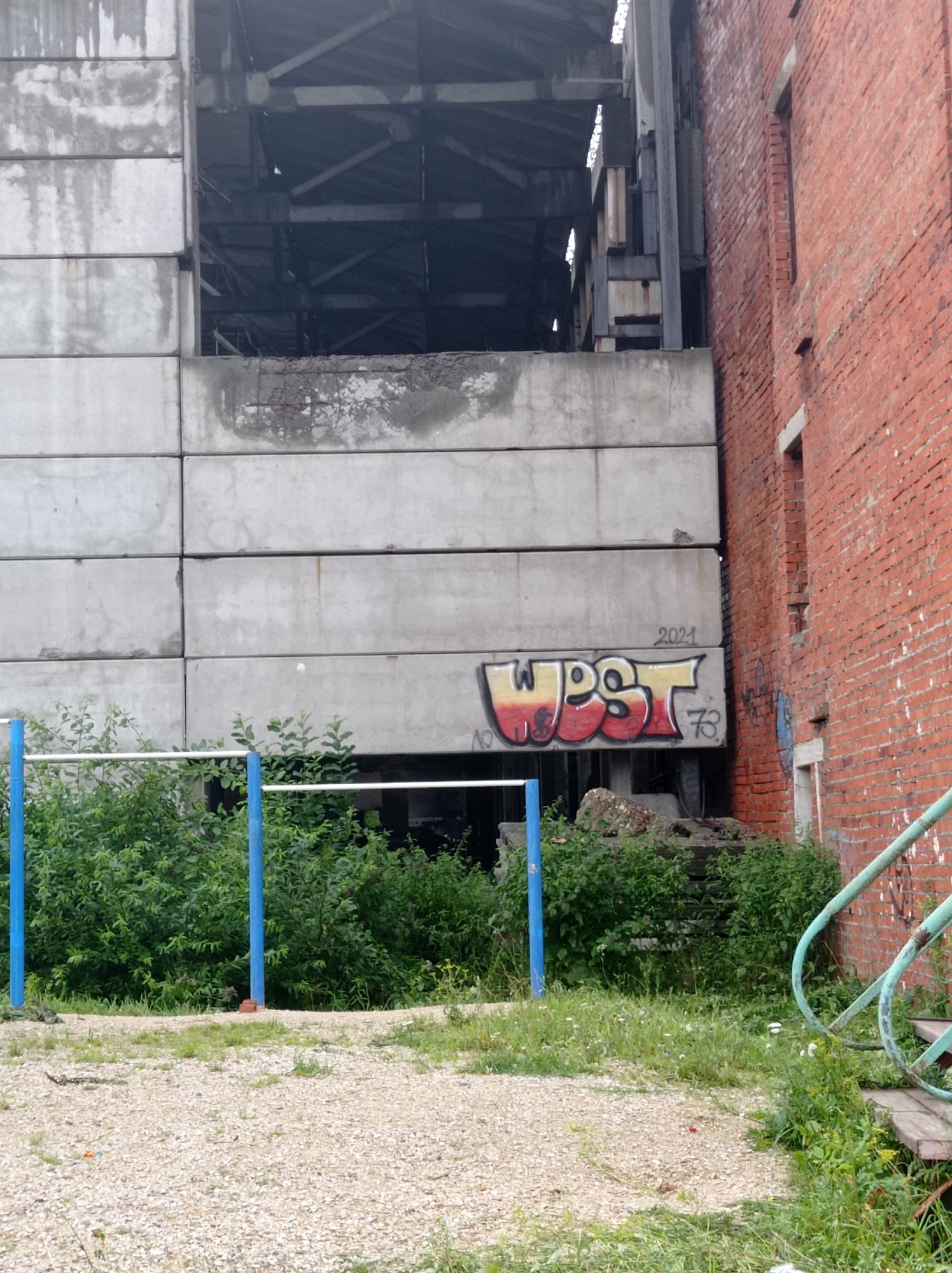 The image depicts an outdoor scene with an unfinished or abandoned industrial building on the left, constructed with large concrete slabs. A red brick building stands to the right. Graffiti is present on the concrete structure, including the word "WPST" in yellow and red, along with the numbers "2021" and "73". In the foreground, a section of a blue and white horizontal bar structure, likely part of an outdoor exercise apparatus, is visible. Lush green vegetation, including trees and bushes, grows around the structure and in front of the buildings. The ground is covered with gravel and sparse grass. The scene appears to be located in an urban or semi-urban area, and based on the context provided about the city of Degtyarsk, Russia, this location is likely within that area. There are no people visible in the image.