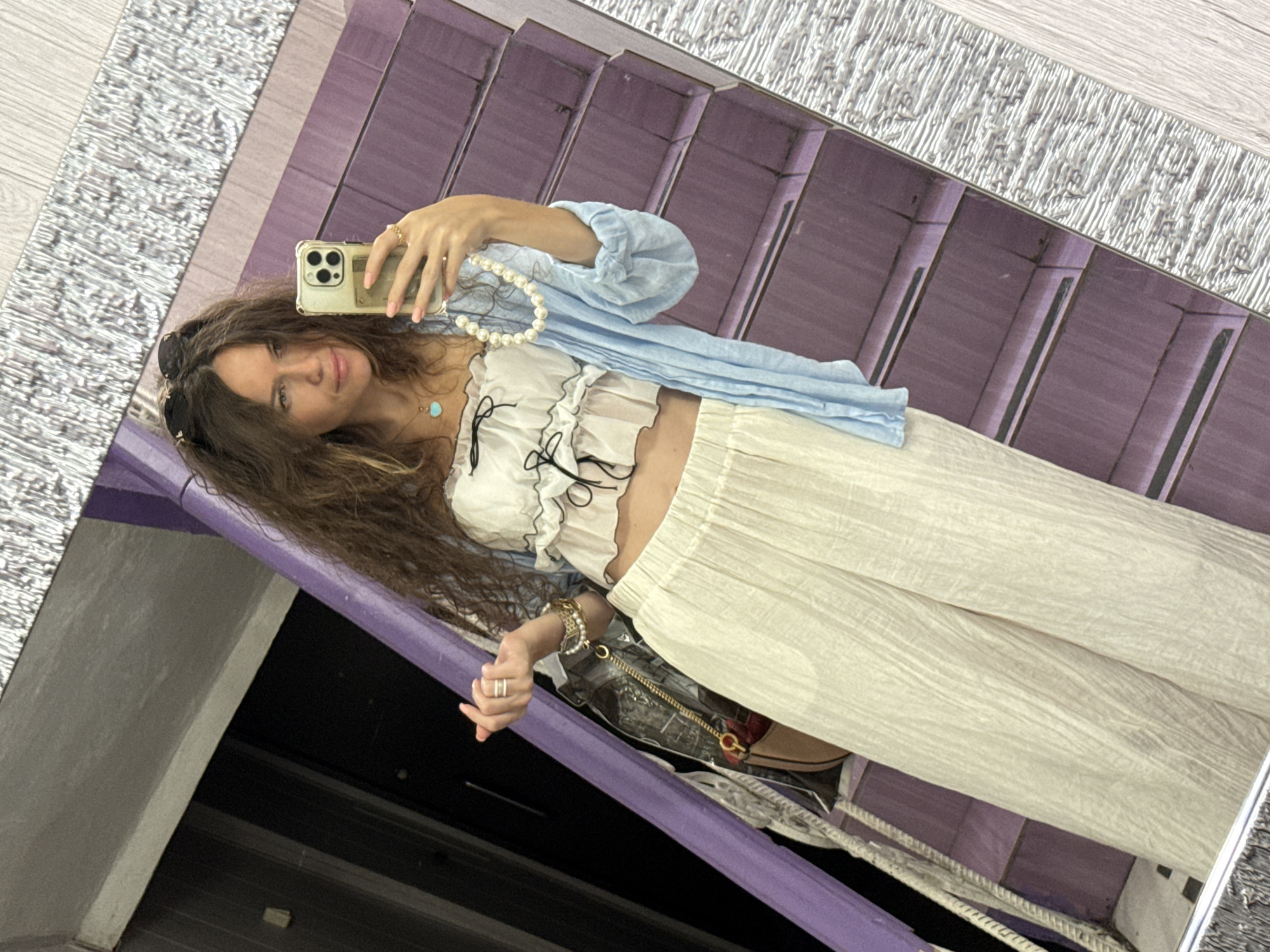A woman is taking a mirror selfie in a hallway with purple stairs. She is wearing a white ruffled crop top, a light blue unbuttoned shirt, and off-white wide-leg pants. She is also wearing a pearl necklace, several bracelets, and rings on her left hand, and a turquoise pendant necklace. A patterned bag is visible on her left side, and she is holding her phone in her right hand. Her hair is long and brown and appears to be naturally wavy. The hallway appears to be indoors, with a dark doorway to the left and a light-colored floor. The purple stairs are reflecting in the mirror, and the overall aesthetic suggests a bohemian or resort style.