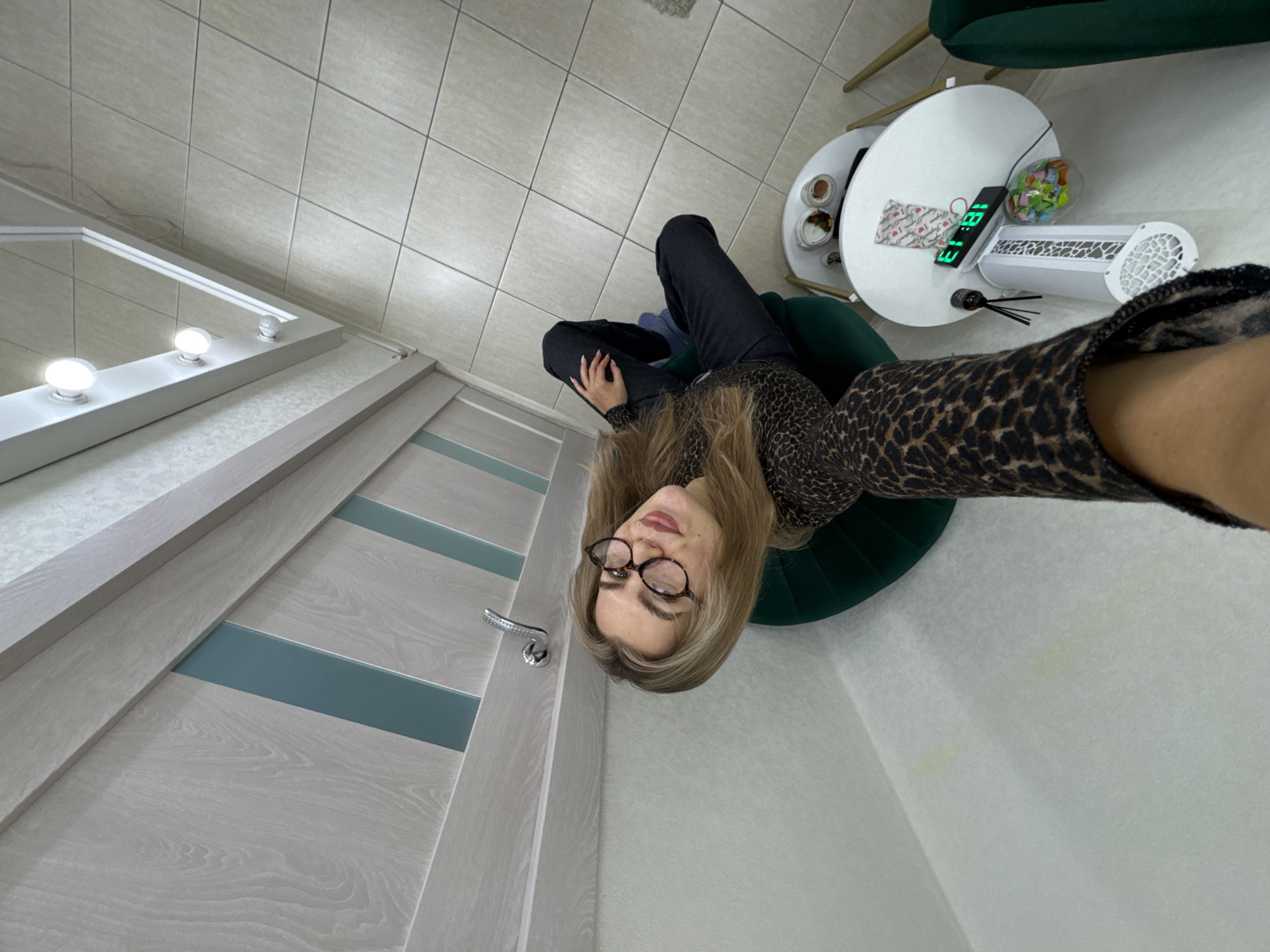 A top-down view of a room featuring a woman wearing glasses and a leopard-print top, seated in a green chair. She's positioned near a door that appears to be ajar and a vanity mirror. Next to her is a small, round white table holding an electronic device displaying the time "18:13", along with other objects. The room has tiled floors, and the lighting appears to be bright, suggesting an indoor environment.