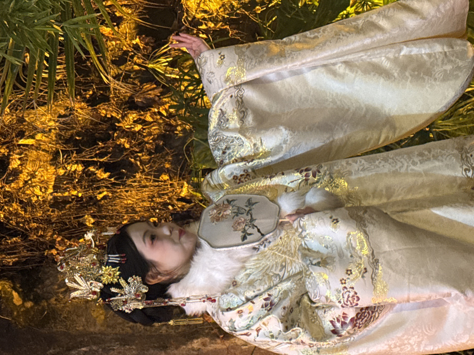 A single East Asian female individual is depicted in an evening outdoor setting within Yuzhong District, China. She is positioned facing slightly right of the camera, head turned, and gaze directed towards the right, exhibiting a serene facial expression. Her dark hair is styled into an elaborate bun, heavily adorned with a prominent golden crown-like piece, intricate jeweled hairpins, and red-beaded dangling ornaments. She wears an opulent, voluminous traditional Chinese garment, primarily light gold or off-white in color, featuring extensive golden embroidery depicting phoenixes and red and maroon floral patterns. A white, fluffy fur collar is visible around her neck. In her left hand, she holds a small, circular, light-colored fan embroidered with a multi-petaled floral motif. Her right arm is extended to the right, with the hand gracefully curved and fingers splayed slightly, palm facing upwards. The immediate background consists of dense, dark green foliage, including tangled vines and slender plant fronds, intensely illuminated by a warm, golden-yellow light, creating a high-contrast environment.