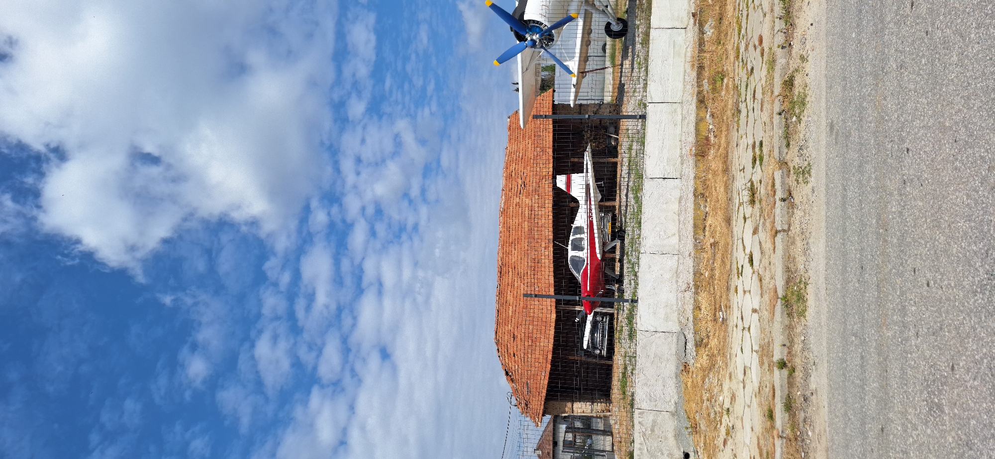 The image depicts two airplanes parked behind a chain-link fence in Kapatovo, Bulgaria. A single-engine red and white airplane is located under a brick-roofed structure. Further along, another white airplane is visible, with blue and yellow propeller blades. The airplanes are near a sidewalk and a paved road. In the background, there is a blue sky with scattered clouds.