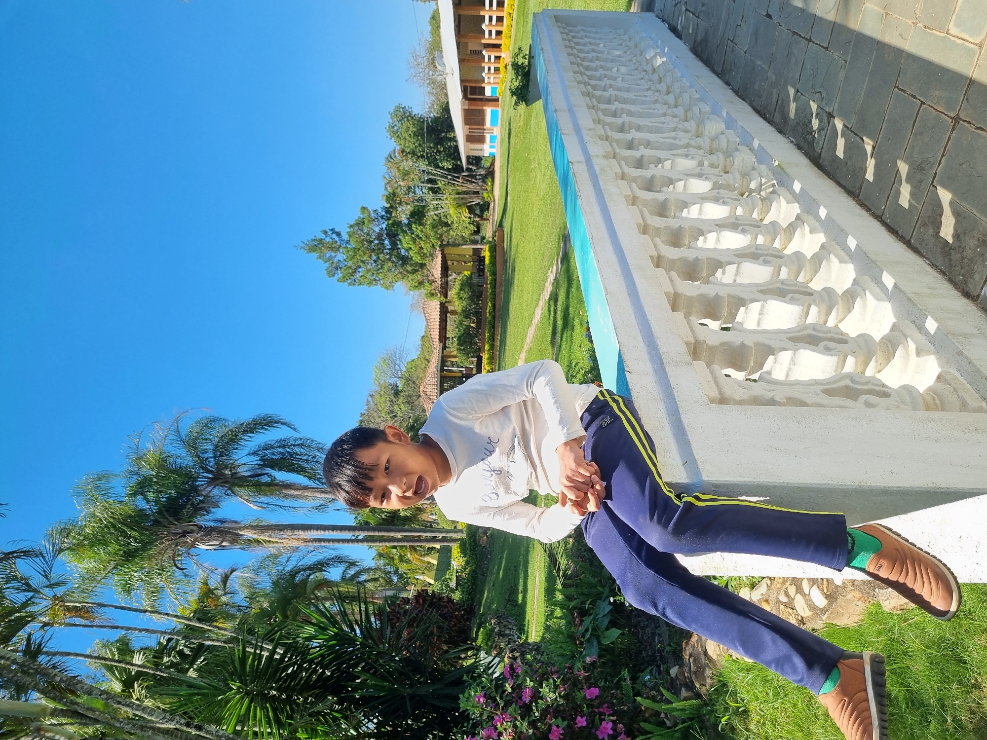 A young Asian boy sits on a white, ornate balustrade outdoors in Laranjal Paulista, Brazil. He wears a long-sleeved white shirt and dark blue athletic pants with a yellow stripe. His feet are clad in brown slip-on shoes and bright green socks. The boy appears to be speaking, with his mouth open and hands clasped loosely in his lap.
The background reveals a lush green lawn dotted with palm trees and other foliage. Several buildings with terracotta roofs are visible in the distance, suggesting a park or resort setting. The sky is a clear, bright blue. The balustrade he sits on is made of white concrete with decorative columns and is positioned next to a stone pathway. The overall scene suggests a warm, sunny day in a tropical or subtropical environment.