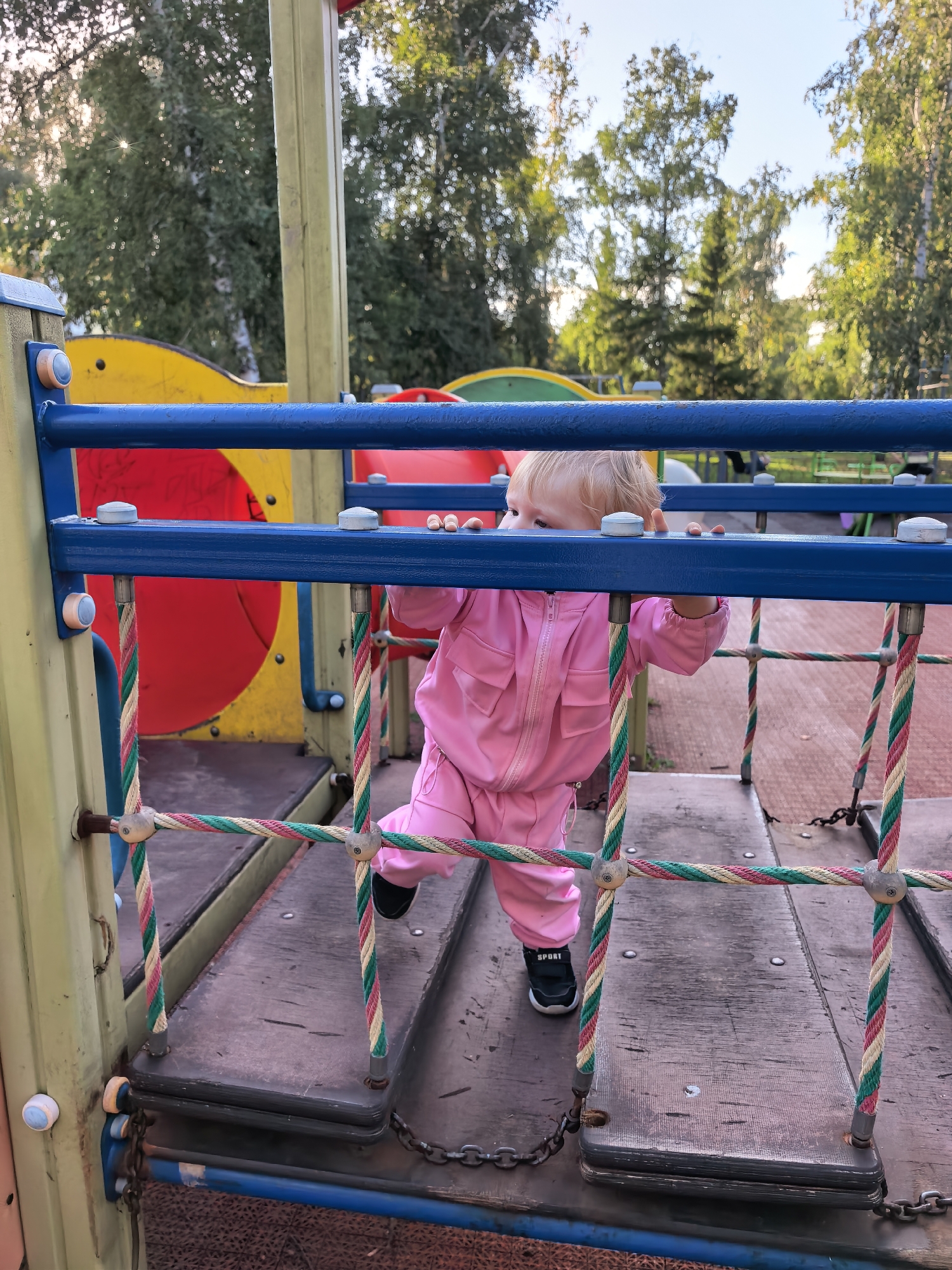 A young child wearing a pink jumpsuit is on a playground structure. The child is positioned in the center of the frame, looking to the left, with their hands resting on a blue horizontal bar. The playground equipment is made of metal and wood, with colorful panels in the background, including red and yellow sections. Ropes with red, green, and white stripes form a net-like structure around the child. The ground beneath the structure appears to be a reddish-brown rubberized surface. Trees and foliage are visible in the background, suggesting an outdoor setting. The lighting indicates it is daytime. The specific location in Sterlitamak, Russia, is not discernible from the image itself.
