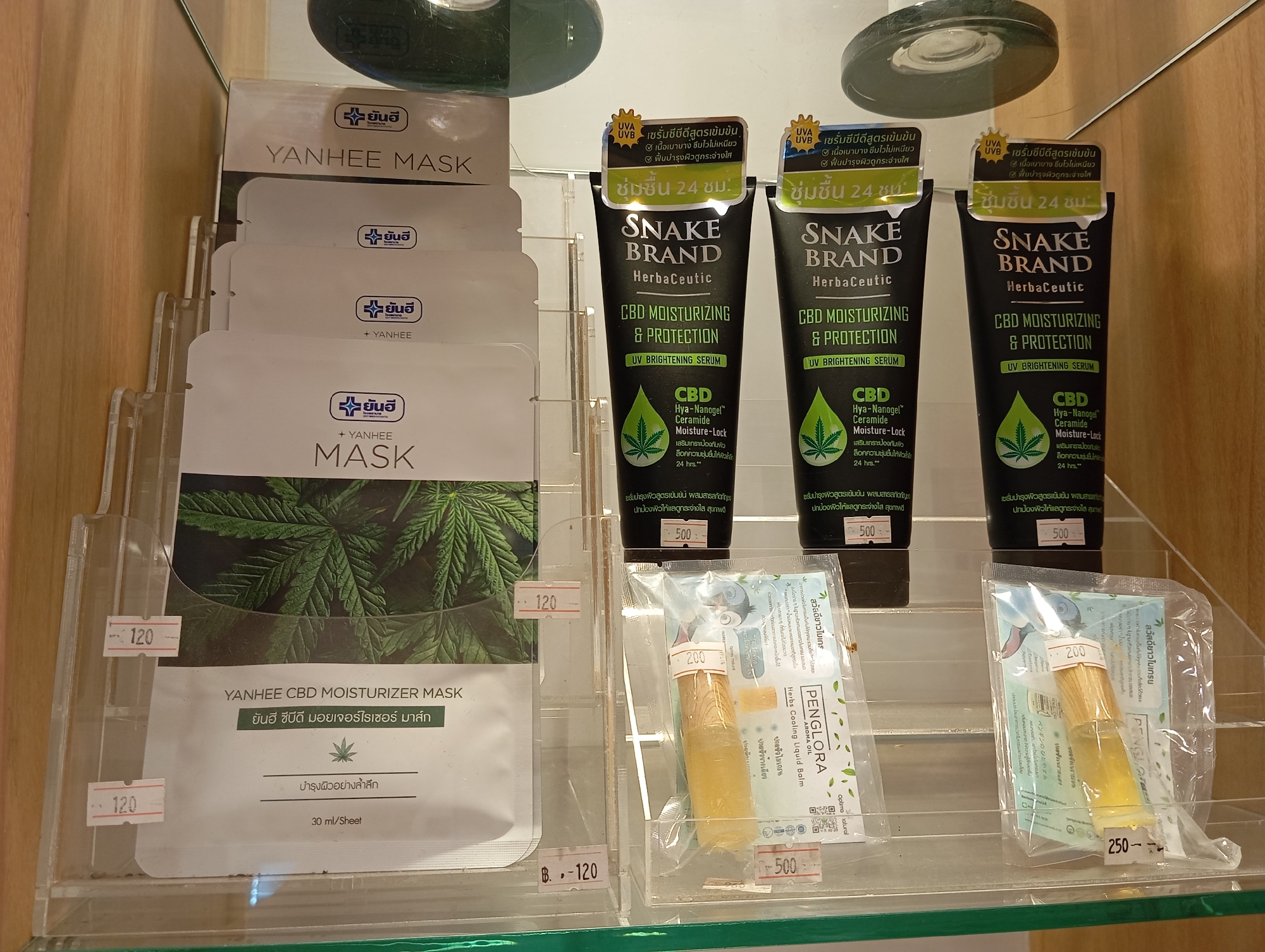 This is a close-up, eye-level shot of a retail display shelf in Karon, Thailand, showcasing skincare products. The display is made of clear acrylic. On the left are several individually packaged facial masks. Each mask package features a prominent logo with a cross and the text " yanhee" in Thai and English. The front-most mask package is white with a green marijuana leaf motif and reads "YANHEE MASK" and "YANHEE CBD MOISTURIZER MASK". Price tags of "120" and "B. 120" are visible on the left side of the mask display.
To the right of the masks are three tubes of "SNAKE BRAND HerbaCeutic CBD MOISTURIZING & PROTECTION UV BRIGHTENING SERUM". These tubes are black with yellow and green labels. One of these tubes is partially obscured by a clear plastic container holding two vials. The labels on the tubes display information in both English and Thai, mentioning "CBD" and a marijuana leaf icon. Price tags of "500" are visible below the tubes.
Further to the right, within a clear plastic container, are two small vials of "PENGLORA Aroma oil Herbs Cooling Liquid Balm". These vials contain a yellow liquid and have wooden stoppers. One of these vials has a price tag of "200" attached to its packaging. Below this, another price tag reads "250". The background consists of a light-colored surface, likely a shelf or wall, and part of a dark, rounded object, possibly a light fixture. The overall scene suggests a commercial environment, likely a pharmacy or a shop selling beauty products.