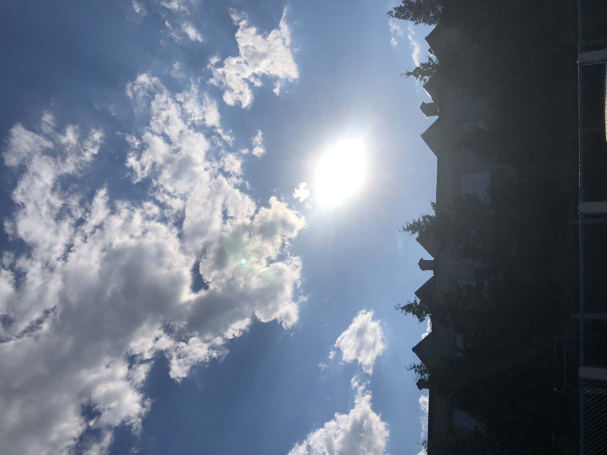 The image shows a bright sun shining through clouds in a blue sky, casting a lens flare in the center of the frame. Below the sky, silhouetted against the bright background, is a row of buildings with peaked roofs and chimneys, partially obscured by trees. A dark fence runs across the bottom of the image. The location is the city of Khmilnytsia, Ukraine. There are no people visible in the image.