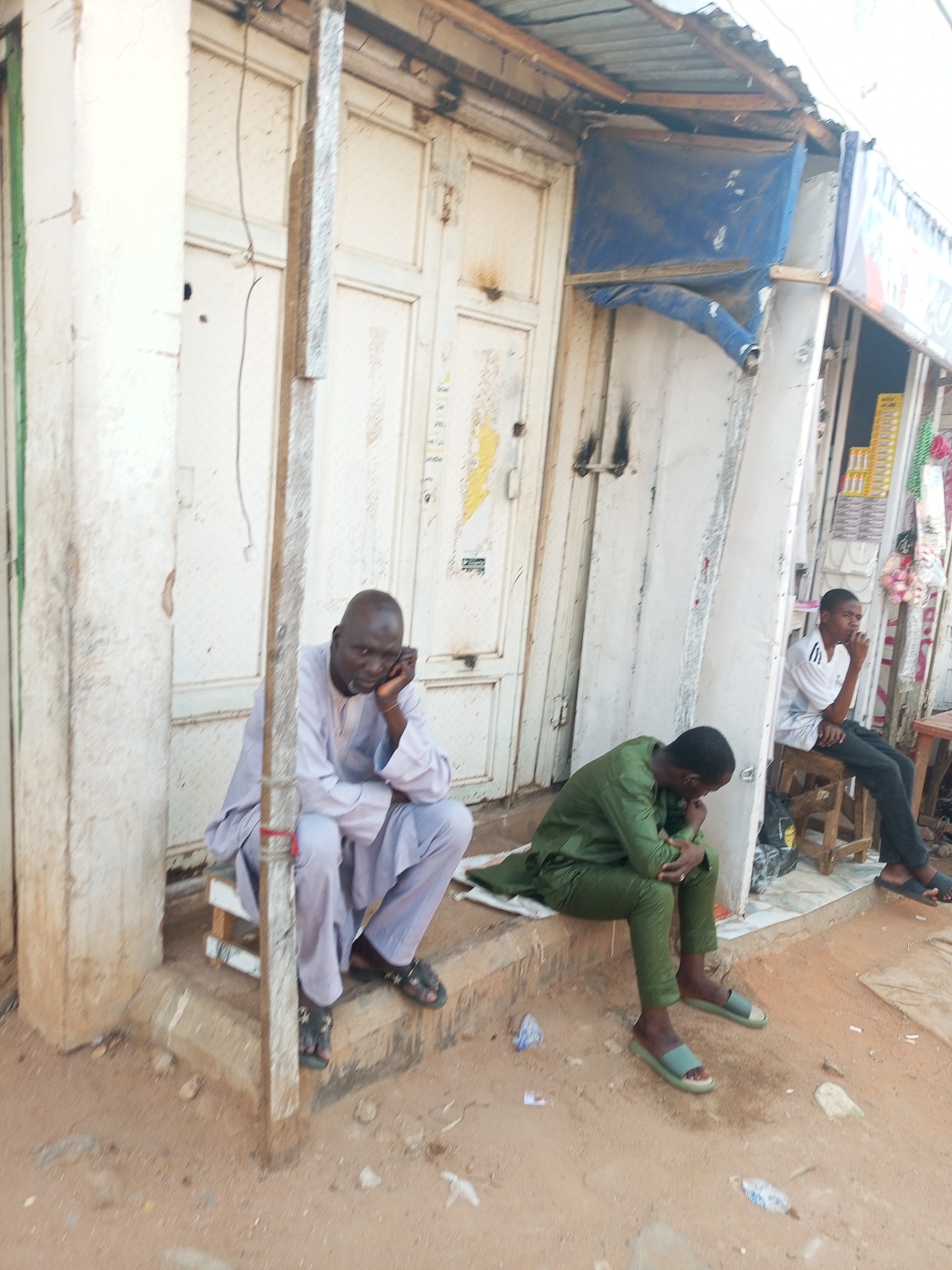 Three men are sitting in front of a building with white doors. One man is wearing a light purple robe and sandals. Another man is wearing a green robe and sandals. The third man is wearing a white shirt with stripes and black pants. The building appears to be a shop with a partially open storefront. The ground is dusty and sandy. The image suggests an urban setting, but lacks clear identifiers for Tempure, Nigeria.