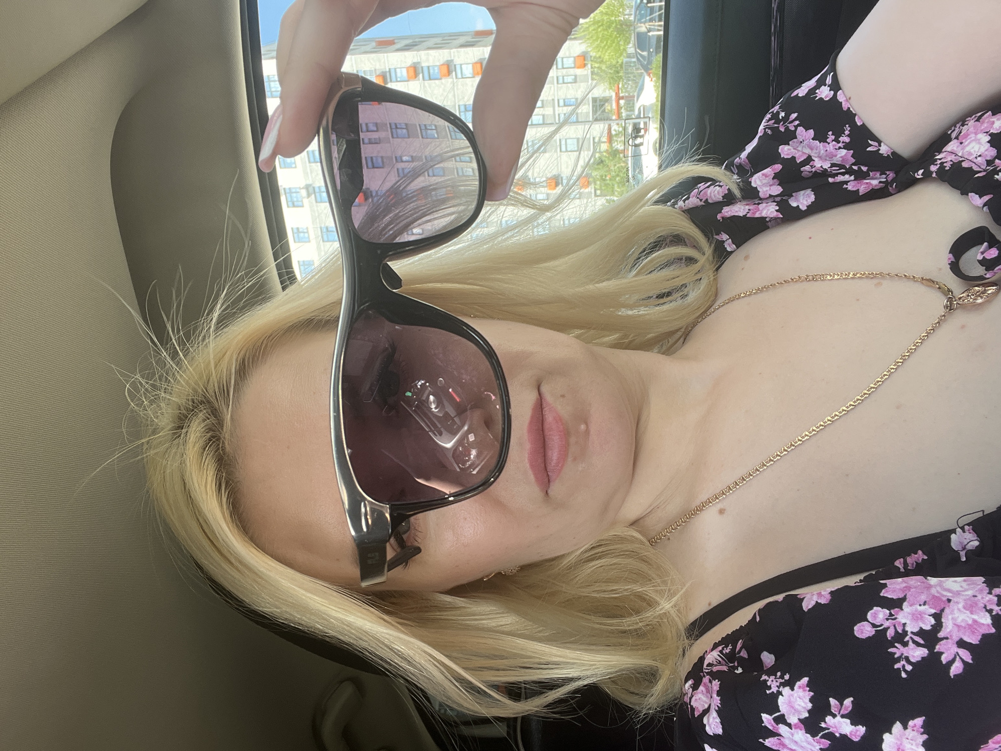A blonde woman in a black floral dress is sitting in a car, holding up a pair of sunglasses to obscure her face. The sunglasses are dark, with a metallic frame. Her left hand, with a light pink manicure, is visible holding the sunglasses. In the reflection of the sunglasses, a car's dashboard and a person's face can be seen. The background, visible through the car window, shows a modern residential building with many windows and balconies, indicative of urban development. Trees and other cars are also visible, suggesting an outdoor setting in a city. The lighting suggests it is daytime, possibly a sunny day. No specific landmarks are identifiable to pinpoint the location within Tyumen, Russia, beyond it being an urban environment.