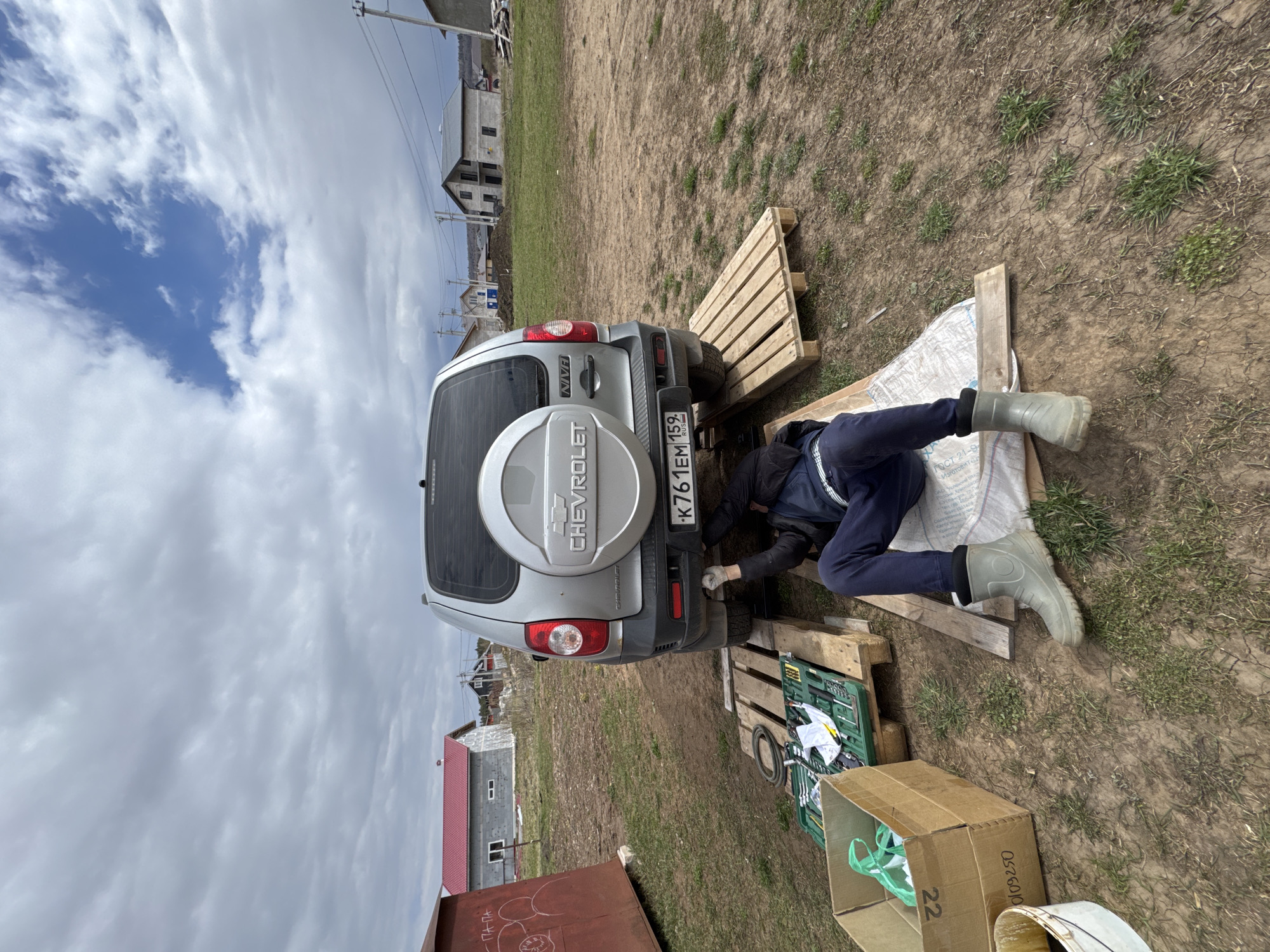 A person lies under the rear of a silver Chevrolet SUV in a yard at Ферма, Russia. The SUV has a spare tire mounted on the rear door and a license plate reading "K761EM 159". The person is lying on a white sheet placed on top of wooden planks and pallets. They wear dark blue pants, a dark jacket, and light-colored boots. A green toolbox and a cardboard box containing miscellaneous items are placed beside the pallets. The yard appears to be dirt and sparse vegetation. A building with a red roof is partially visible in the background. Other buildings and utility poles are visible further in the distance under a cloudy sky.