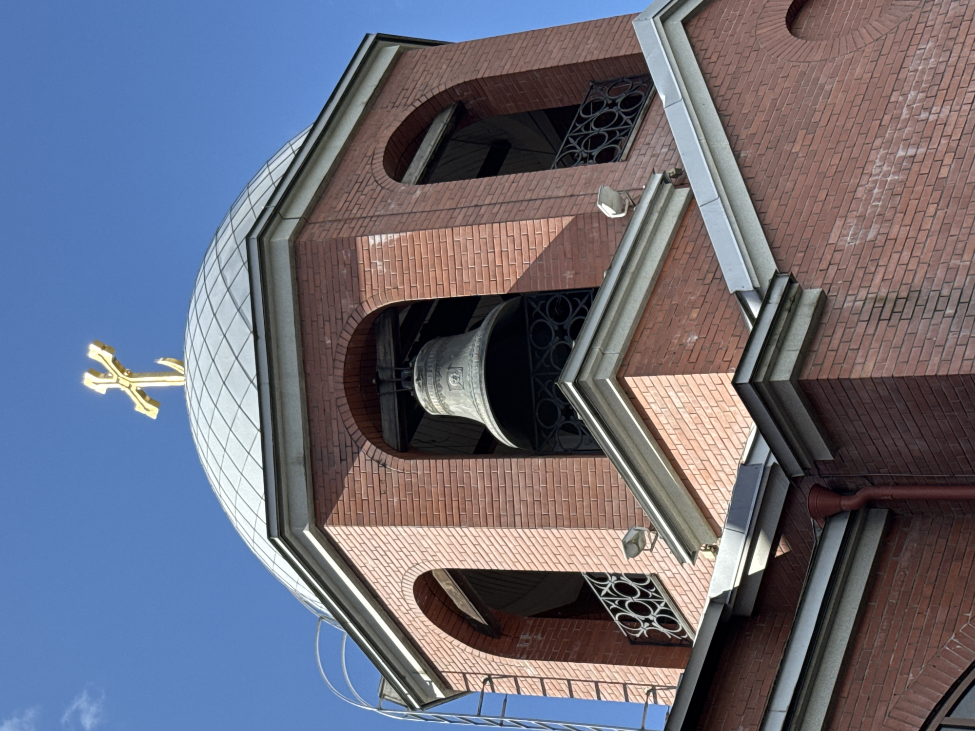 The image shows the top of a brick bell tower of a church in Saint Petersburg, Russia, under a clear blue sky. A large, dark bell hangs within an arched opening of the tower. Above the bell tower, a silver-colored dome is topped with a golden cross. The architecture suggests an Orthodox Christian church, common in Saint Petersburg. The brickwork is red and the dome is paneled. There are decorative metal railings on the openings of the tower and a light fixture is mounted on the exterior wall. A metal ladder runs up the side of the tower. The scene is a detail shot, focusing on the upper portion of the structure.