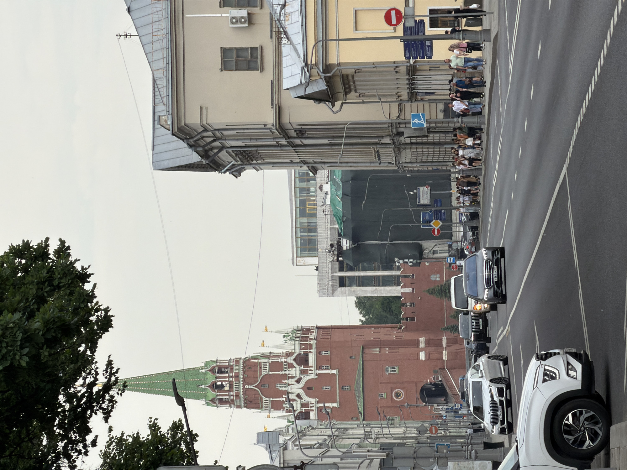 A street scene in Moscow, Russia, features the iconic Spasskaya Tower of the Kremlin in the background, identifiable by its red brick construction, green spire, and white decorative elements. Several cars are on the road, including a white sedan, a dark SUV, and a dark gray sedan. A white SUV is parked on the left side of the frame. Pedestrians are walking on the sidewalk to the right of the street, and a few are visible in the background near the Kremlin. Buildings line the street on the right, with architectural details typical of historic city centers. Streetlights and traffic signs are present. The sky is overcast, suggesting a cloudy day. The overall atmosphere is of a busy urban environment with a prominent historical landmark.
