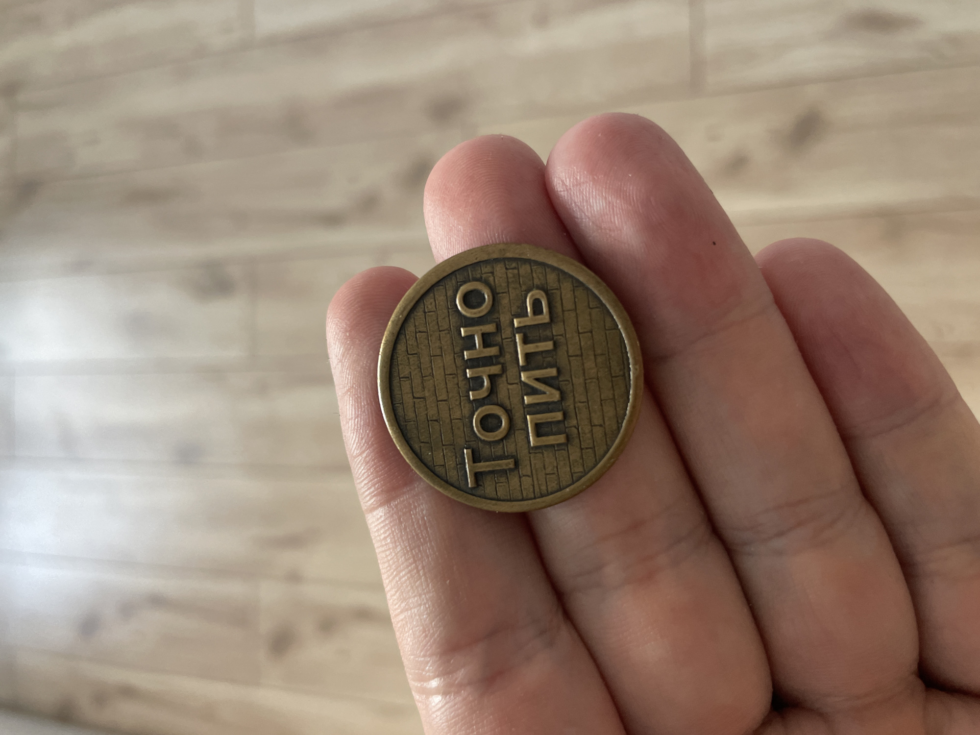 A close-up, eye-level shot shows a person's hand holding a round, bronze-colored coin. The coin features raised lettering that reads "ТОЧНО ПИТЬ" against a brick-like background texture. The coin is positioned between the thumb and index finger, with the palm of the hand visible supporting it from below. The background is a light-colored wooden floor.