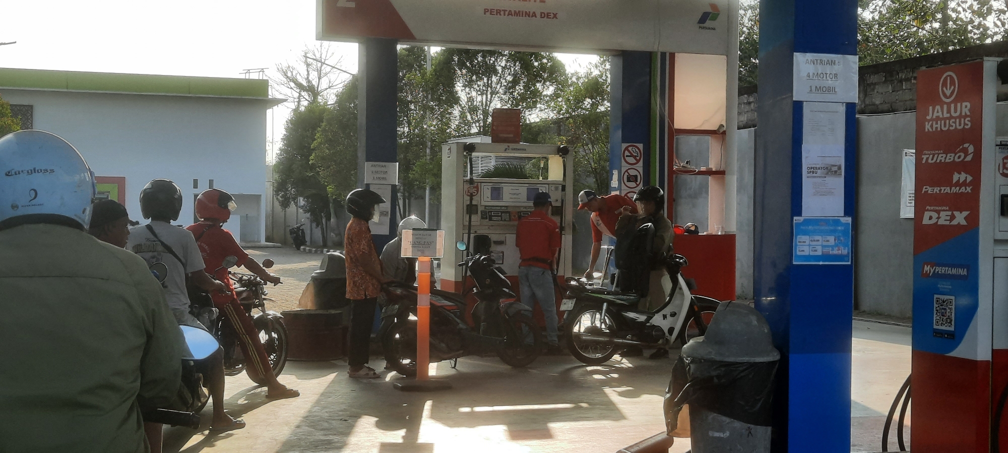 This is a medium-shot, eye-level, outdoor photograph taken at a Pertamina gas station in Lodoyo, Indonesia. The scene depicts a typical gas station environment with several motorcycles and people. The gas station structure is visible, including the fuel pumps and the overhead canopy. The lighting suggests it is daytime, possibly late afternoon, with shadows cast on the ground.
In the foreground, on the left, a person wearing a green jacket and a blue helmet is partially visible, facing away from the camera. To their right, a person wearing a black helmet is on a motorcycle. Further to the right, another person wearing a red helmet is also on a motorcycle.
In the middle ground, near the fuel pumps, several individuals are interacting. One person in an orange and black patterned shirt and dark pants is standing and looking down. Another person in a gray shirt and white cap is seated on a motorcycle. Two gas station attendants are also present; one in a red shirt and blue jeans, and another in a red shirt and a white cap, appearing to be assisting customers. Several motorcycles are parked and in use at the pumps.
The background shows trees, a white building with a green trim, and a gray brick wall. Signs are visible on the gas station pumps, indicating fuel types and queuing instructions. One sign reads "ANTRIAN: 4 MOTOR 1 MOBIL," indicating the queue order. Another sign has the Pertamina logo and various fuel brand names like "TURBO," "PERTAMAX," and "DEX." A trash can with a black trash bag is situated on the right side of the frame. The ground is paved concrete, with visible shadows from the structures and people.