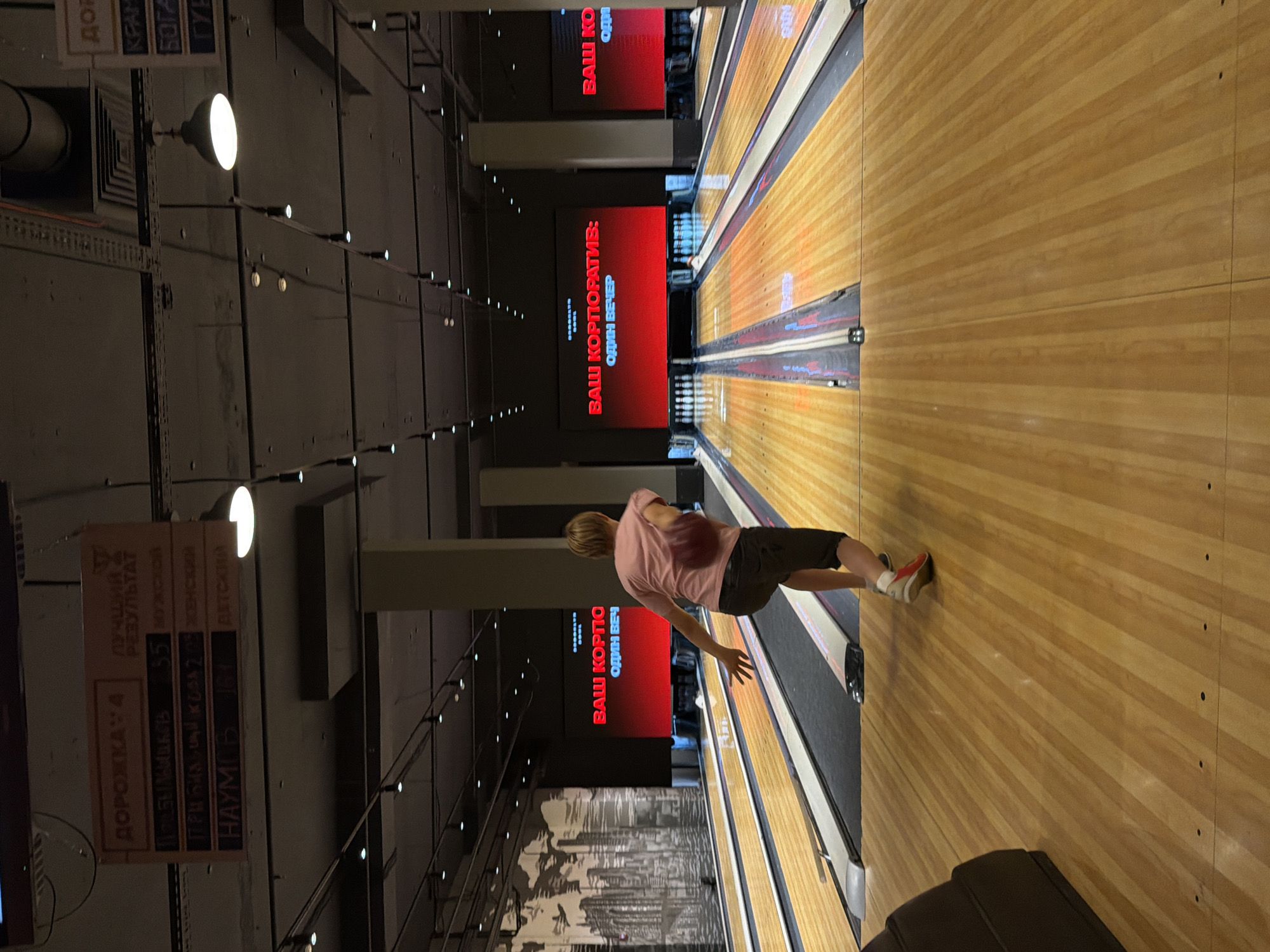 The image depicts a bowling alley in Izhevsk, Russia. The primary subject is a young person, likely male, mid-action, throwing a bowling ball down a lane. He is wearing a pink t-shirt and dark shorts, with athletic shoes. The bowling alley has wooden lanes, typical for the sport. Above the lanes, large red screens display text in Russian, reading "ВАШ КОРПОРАТИВ: ОДИН ВЕЧЕР" which translates to "YOUR CORPORATE EVENT: ONE EVENING". This suggests the venue is hosting corporate events.
To the left of the bowling lanes, there is a wall with industrial-style elements and lighting fixtures. Hanging signs provide information, including "ДОРОЖКА 4" (Lane 4) and what appears to be a scoreboard or ranking system with names and scores. One sign lists "ЛУЧШИЙ РЕЗУЛЬТАТ" (Best Result) and numbers like 55 and 161. Another sign seems to indicate categories such as "МУЖСКОЙ" (Male), "ЖЕНСКИЙ" (Female), and "ДЕТСКИЙ" (Children's).
The overall lighting suggests it is likely evening or indoors, with artificial lighting illuminating the scene. The atmosphere appears casual and recreational, focused on the activity of bowling. The perspective of the image is slightly tilted, adding a dynamic feel to the composition.