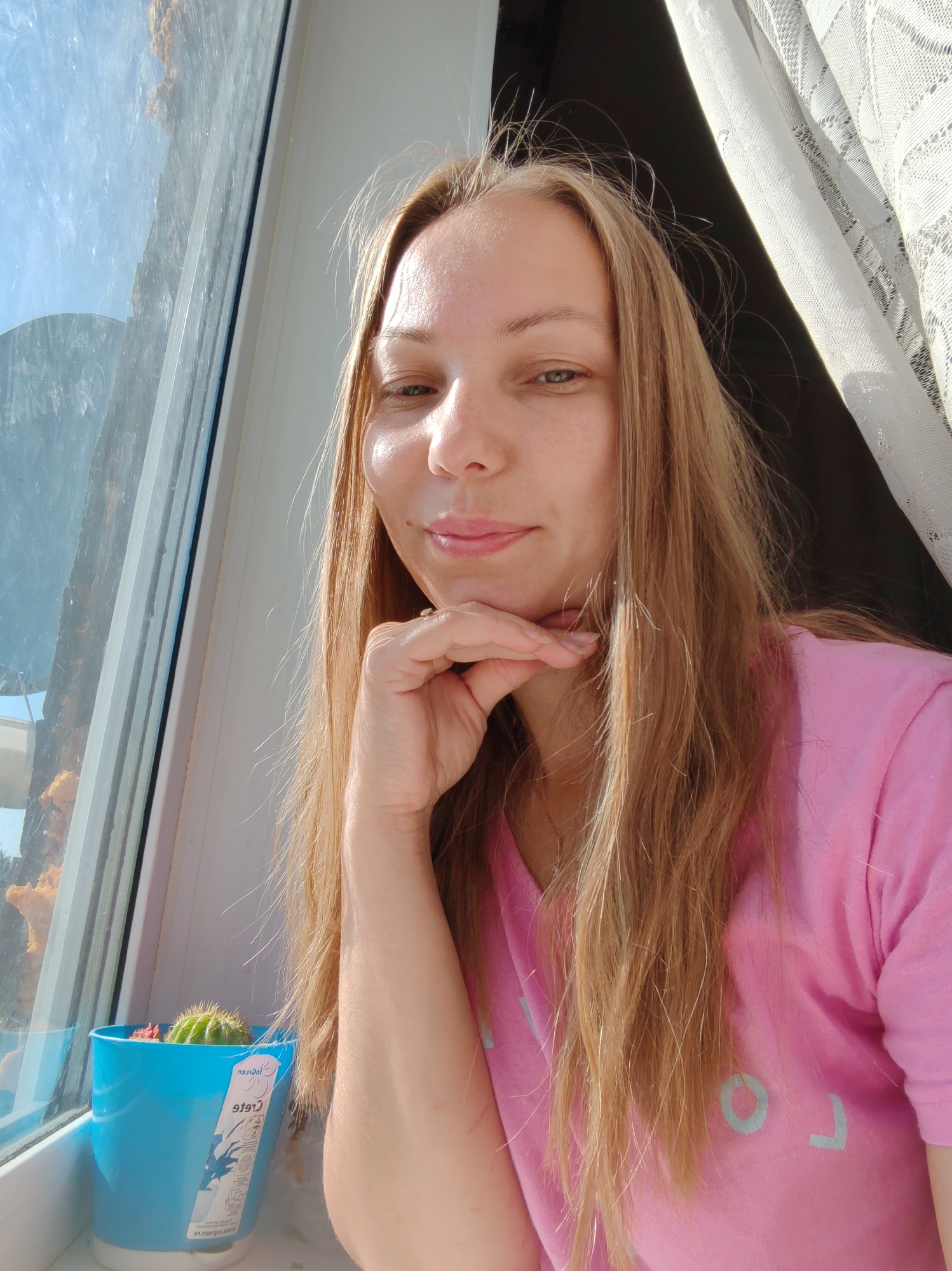 A young woman with long, blonde hair, wearing a pink t-shirt, poses for a selfie next to a window. Her hand is resting on her chin, and she is looking towards the camera with a slight smile. To her left, a small cactus sits in a bright blue pot on the windowsill. The window offers a view of the sky and some distant structures. A lace curtain is visible on the right side of the image. The scene appears to be indoors, near a window.