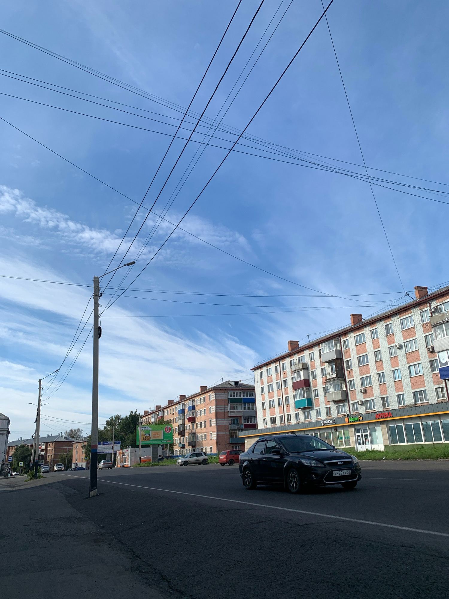 The image depicts a street scene in Prokopyevsk, Russia. A black Ford Focus sedan is driving on the right side of the road. In the background, there are multi-story brick apartment buildings with numerous windows and balconies. Overhead, a complex network of electrical wires stretches across the bright blue sky, which is partially filled with wispy white clouds. A concrete utility pole with attached streetlights stands on the left side of the road. The road itself is paved and appears to be a multi-lane street with white lane markings. The general environment is urban, characterized by buildings, road infrastructure, and utility lines. There are no visible people in the frame.