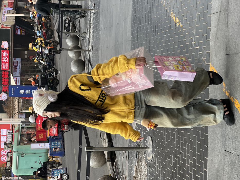 A young woman stands on a sidewalk, wearing a yellow sweater, baggy jeans, and black shoes. She has long dark hair and a white knit cap with headphones. She holds two shopping bags, one pink with cartoon characters and the other transparent, containing a plush toy. The street behind her is lined with scooters, shops with Chinese characters on signs, and a few people. A metal barrier is partially visible in the foreground.