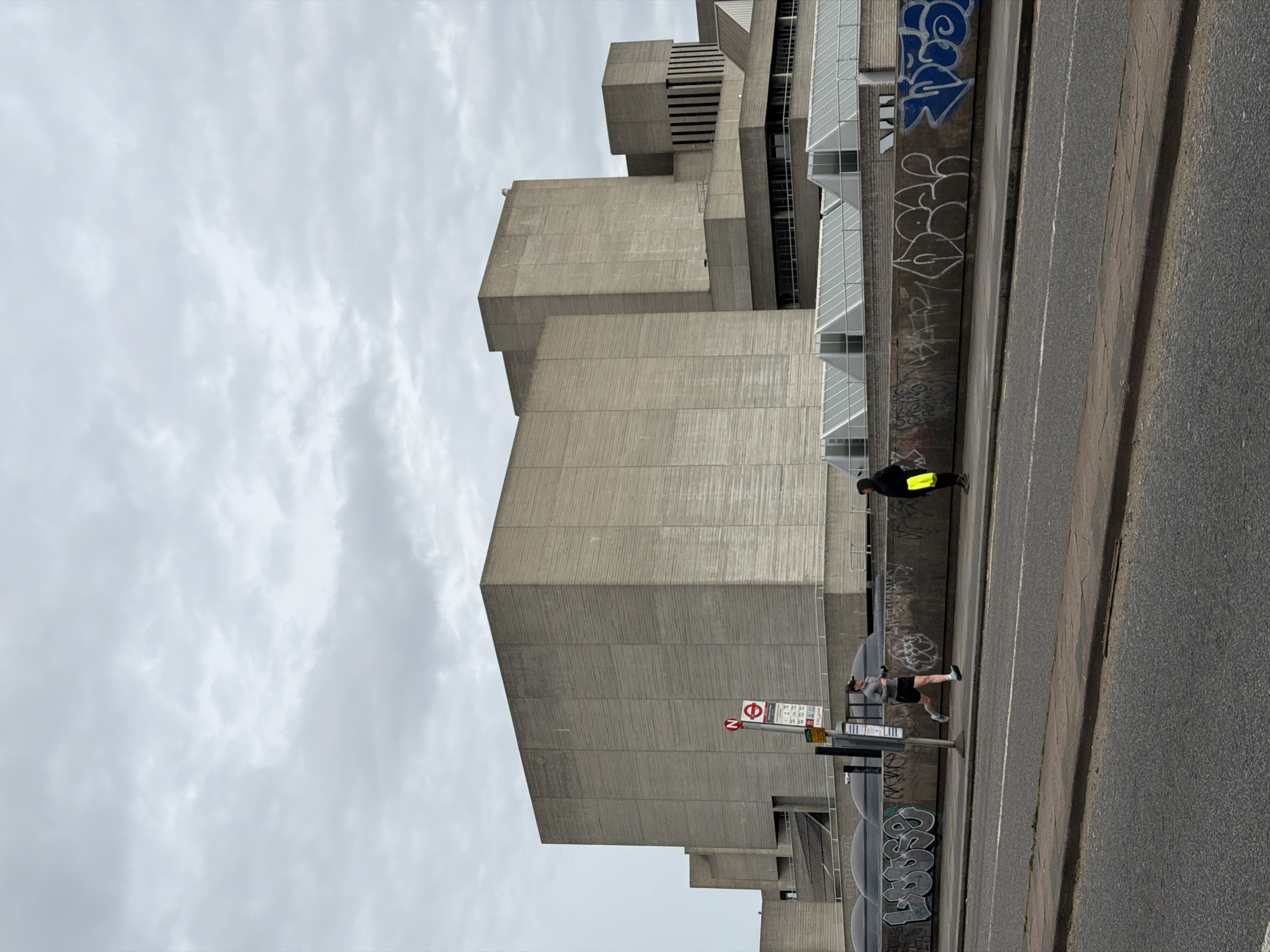 Here's a description of the image:
The photo depicts a cityscape in London, United Kingdom. In the foreground is an asphalt road and a sidewalk along with an edge. A building featuring brutalist architecture is on the left. The concrete exterior of the structure is marked by modular geometric forms and textured surfaces. Graffiti adorns the concrete walls near the street level. People are visible on the sidewalk. One is wearing a black jacket with a bright yellow bag. A bus stop sign is visible along the sidewalk. The sky above is overcast.