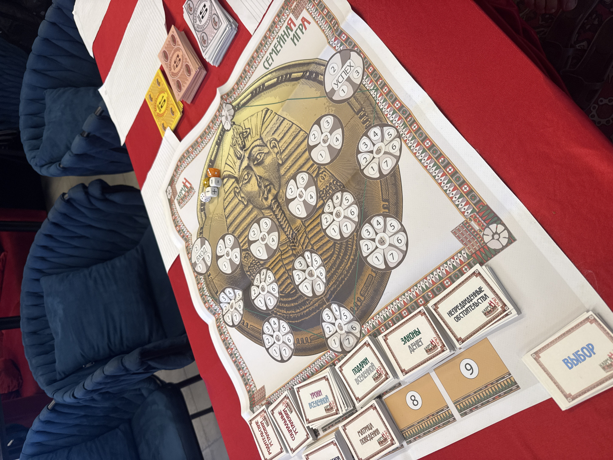 The image displays a vibrant board game setup on a red tablecloth, suggesting an indoor recreational setting. Two dark blue, plush armchairs are visible in the background to the left.
The central subject is a large game board, prominently featuring a stylized ancient Egyptian pharaoh's head (resembling Tutankhamun) in gold and brown tones. The board is titled "СЕМЕЙНАЯ ИГРА" (Family Game) in Cyrillic at the top. Numerous circular segments, each divided into smaller numbered sections (typically 1-7), are scattered across the board. Some segments are labeled "ВХОД" (Entrance) and one prominent segment reads "УСПЕХ" (Success), indicating progression points or objectives within the game. Two dice, one white and one orange, are placed near the pharaoh's head.
Around the board's periphery and on the table, several stacks of game cards are arranged. These cards are labeled in Cyrillic, revealing various thematic elements of the game. Visible card titles include: "РОДИТЕЛЬСКИЕ УСТАНОВКИ" (Parental Settings/Attitudes), "СОЦИАЛЬНЫЕ УСТАНОВКИ" (Social Settings/Attitudes), "УРОКИ ВСЕЛЕННОЙ" (Lessons of the Universe), "МАТРИЦА ПОВЕДЕНИЯ" (Matrix of Behavior), "ПОДАРКИ ВСЕЛЕННОЙ" (Gifts of the Universe), "ЗАКОНЫ ДЕНЕГ" (Laws of Money), "НЕПРЕДВИДЕННЫЕ ОБСТОЯТЕЛЬСТВА" (Unforeseen Circumstances), and "ВЫБОР" (Choice). Several cards also feature the word "КАРМАН" or "КАРМЫ" (Karma), possibly a brand name or game concept. Additionally, stacks of playing cards or game money with denominations "10" and "50" (pink and yellow) and other numbered cards "8" and "9" (yellow/brown) are present.
The overall scene captures a board game ready for play, indicating an imminent or ongoing game session. The detailed components suggest a complex game potentially involving life choices, cosmic lessons, and financial aspects, all within an ancient Egyptian aesthetic.