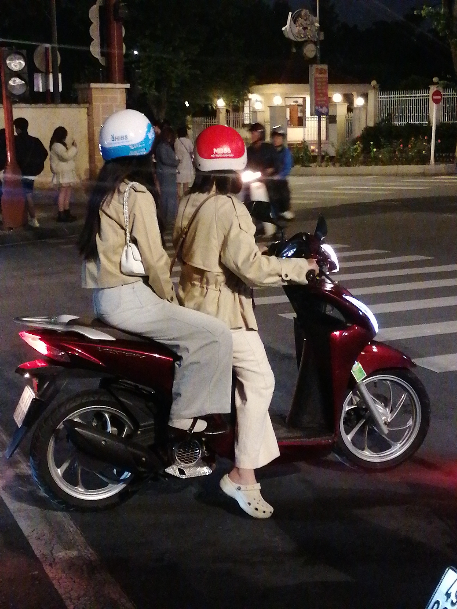 The image shows two individuals riding a dark red scooter at night in Da Lat, Vietnam. The person in the front is wearing a white and blue helmet, and the person in the back is wearing a red helmet. Both are dressed in light-colored jackets and pants. The scooter is on a pedestrian crossing. In the background, there is a building with lights on, a white fence, and trees. The overall scene is illuminated by streetlights and the scooter's headlights.