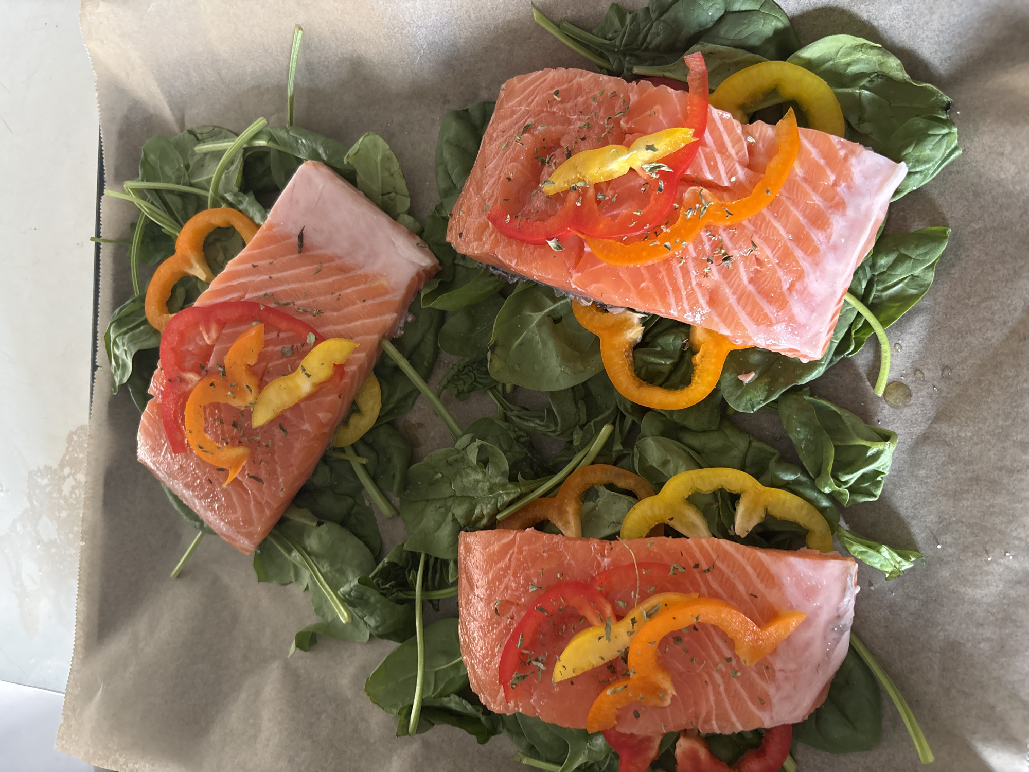 Three raw salmon fillets are placed on a bed of fresh spinach leaves on a baking sheet lined with parchment paper. The salmon fillets are topped with slices of red, orange, and yellow bell peppers, and sprinkled with dried herbs. The scene is photographed from directly above. The background appears to be a light-colored countertop.