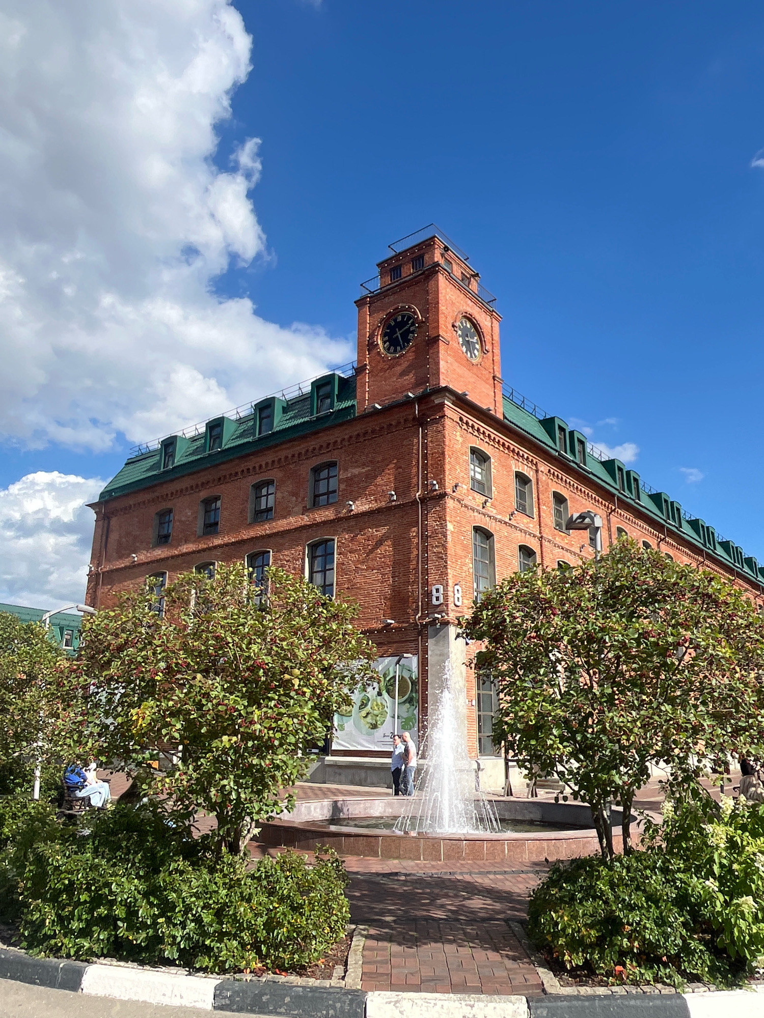 The image shows a red brick building with a green roof and a clock tower, located in Moscow, Russia. The building has multiple windows and dormers on the roof. In front of the building, there is a fountain with water spraying upwards. Several trees with red berries are planted around the fountain. Two people are visible near the fountain, one standing and one sitting on a bench. The sky is blue with white clouds. The scene appears to be an outdoor public space in a city.