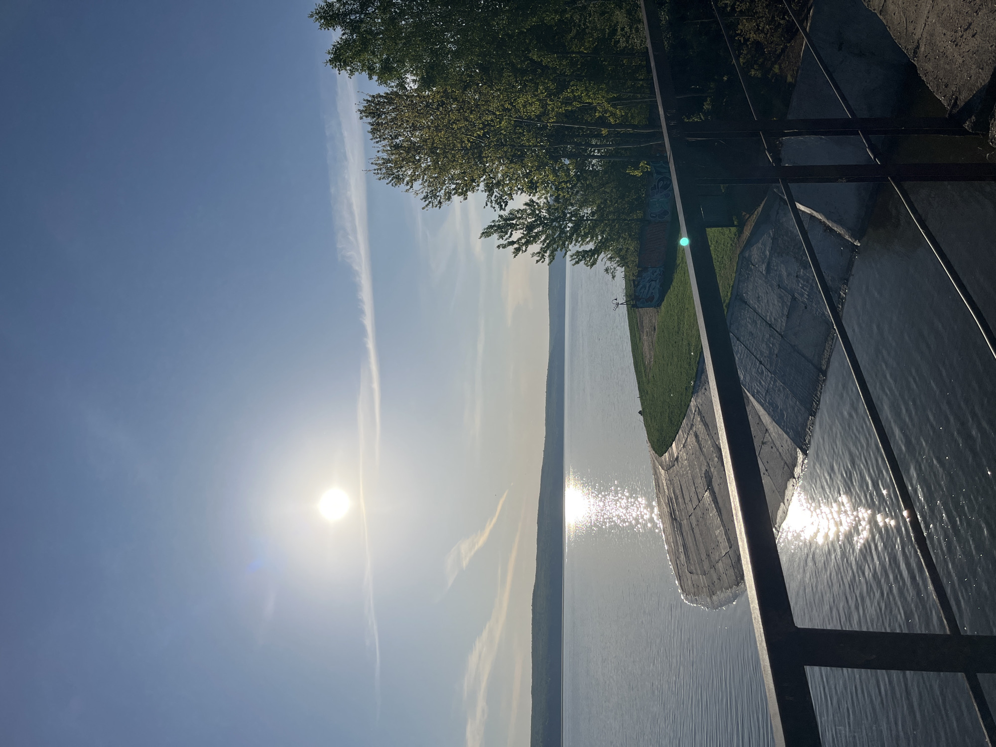 The image shows a view of the Kama River in Chaikovsky, Russia, on a sunny day. The sun is visible in the sky, reflecting brightly on the water's surface. A tree line is visible across the river. On the right of the frame, there is a concrete embankment and a metal railing. Some graffiti is visible on the concrete near the railing.