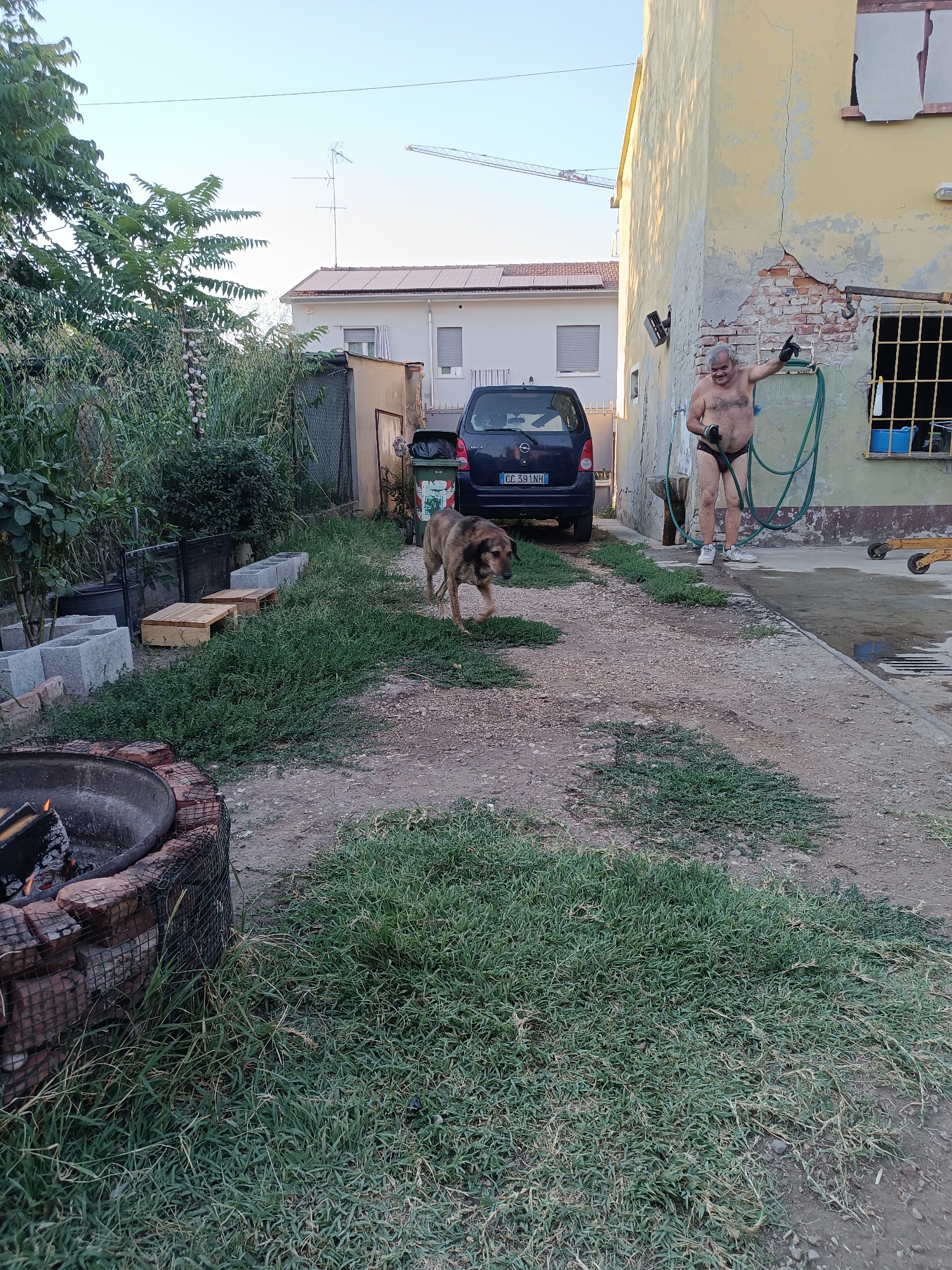 The image depicts an outdoor scene at dusk in Viadana, Italy. In the foreground, a fire pit with burning wood is visible on the left. To the right of the fire pit, a dog walks on a gravel path. In the background, a man is standing outside a yellow building, wearing only black underwear and white sneakers. He is holding a green hose and appears to be spraying water. A dark blue car is parked behind the dog. The environment includes a fence, overgrown vegetation, and other buildings in the distance, including one with solar panels on its roof. A construction crane is also visible in the sky. The overall setting appears to be a residential or industrial yard.
