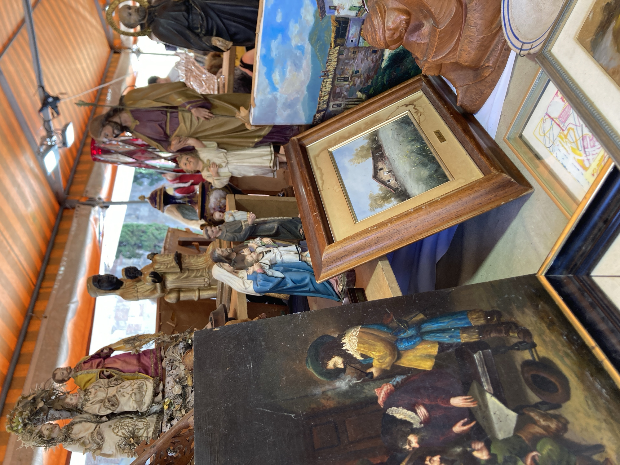The image shows an outdoor market stall under an orange striped awning. The stall displays a collection of religious statues and paintings. In the foreground, a dark-colored oil painting depicts several men gathered around a table, with one man smoking a pipe. To the right, a framed landscape painting shows a rustic building in a grassy field under a cloudy sky. Further back, a large statue of Jesus with a child stands next to a statue of a robed figure with a halo. Other religious figures, including a statue of Mary and child, are also visible. The overall impression is of a religious market, possibly a flea market or a santuario (shrine) market, selling religious artifacts. The context of Barcelona, Spain, is not explicitly visible within the image itself.