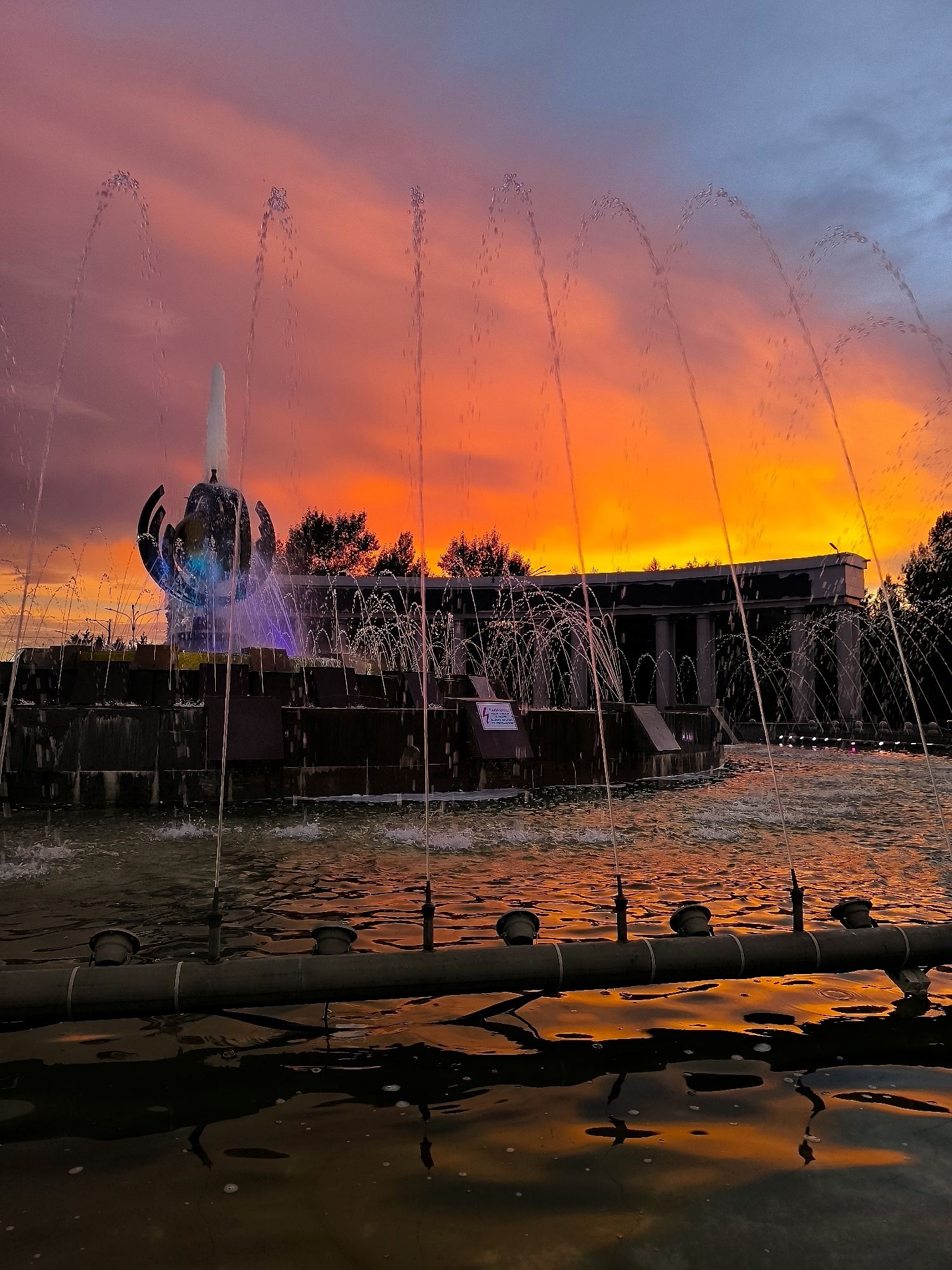 The image captures a vibrant sunset over a fountain in Qarağandı city administration, Kazakhstan. The sky is ablaze with fiery oranges and pinks, transitioning to a dusky blue at the top, creating a dramatic backdrop.
The main subject is a large, multi-tiered fountain with water jets arcing gracefully into the air. In the center of the fountain is a unique sculptural element resembling an open flower or a blossoming bud, illuminated with blue and purple lights. The water reflects the warm hues of the sunset, creating a shimmering, dynamic surface.
In the background, a grand, classical-style building with columns is visible, partially obscured by trees. The overall scene evokes a sense of awe and tranquility, amplified by the intense colors of the sunset. The time of day is clearly evening, as indicated by the sunset. The weather appears clear, allowing for the spectacular display of colors in the sky.
A small sign with text is visible on the left side of the fountain's base, but the text is too small and indistinct to be read. Another sign is also visible on a pillar in the background, also unreadable. The foreground features a dark pipe with nozzles, likely part of the fountain's mechanism, partially submerged in the water and reflecting the colorful sky. The overall mood is one of natural beauty and architectural grandeur at the close of day.