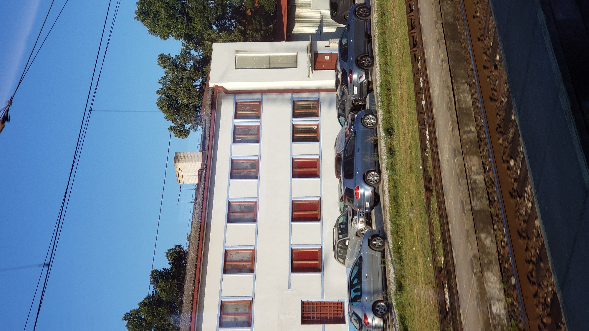 A long shot shows a white industrial building with blue trim and numerous windows with red frames. A parking lot in front of the building is filled with several cars. The building is set back from a train track and a grassy area. Trees are visible behind the building and to the right. The sky is clear and blue.