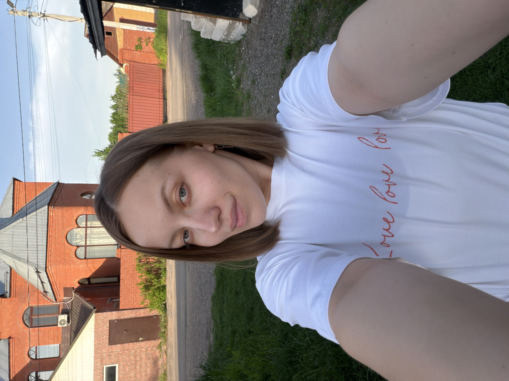 A woman is taking a selfie outdoors. She has light brown hair and is wearing a white t-shirt with a red inscription. Behind her is a red brick house with arched windows. To the right of the house is a patch of green grass and a paved road. The location is likely in Reshetnikovo, Russia based on the prompt.