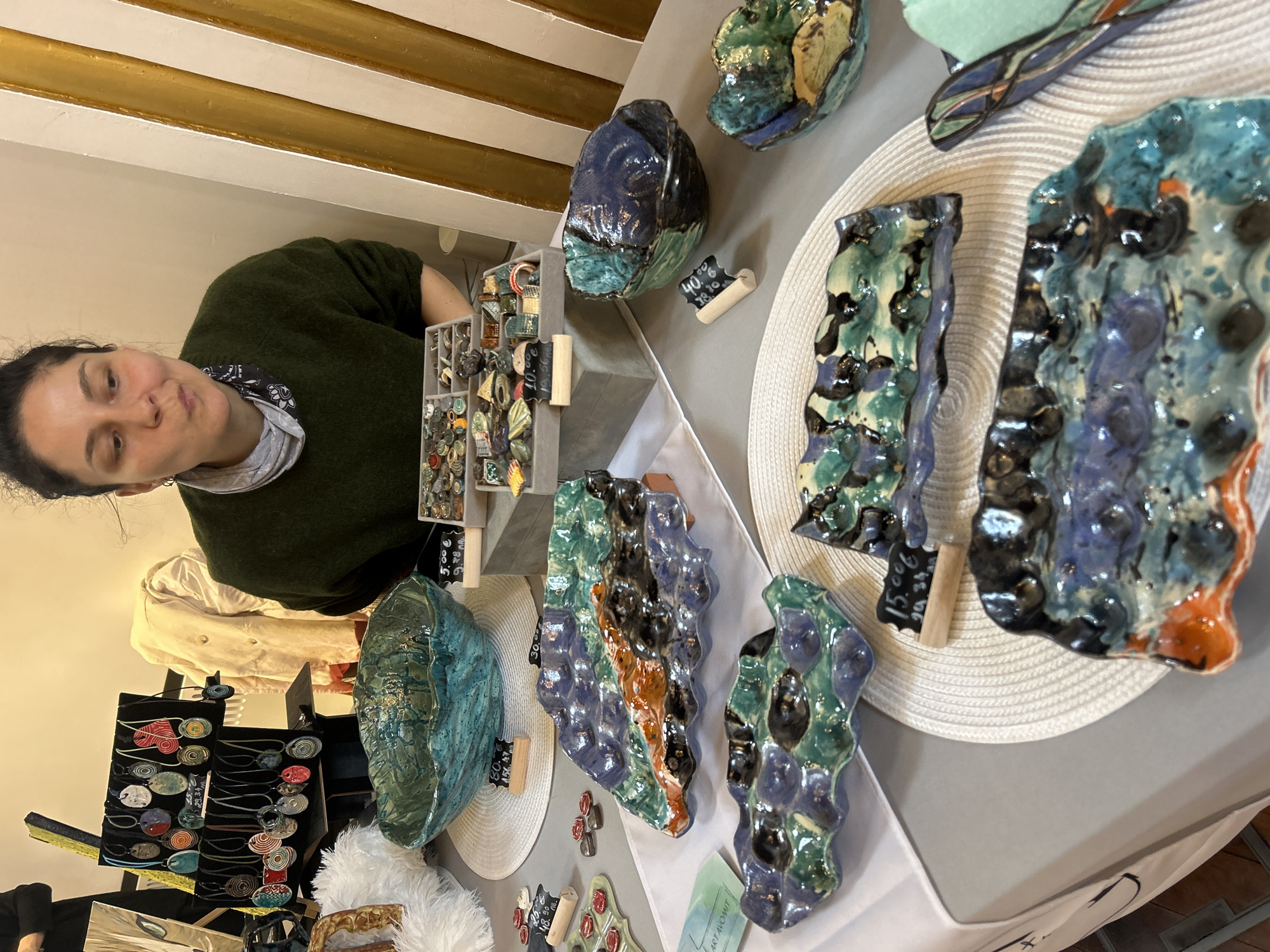 The image shows a woman standing behind a table displaying a variety of handcrafted ceramic items. The items include plates, bowls, and possibly decorative pieces, all featuring unique and colorful glazing. Price tags are visible on some of the items. The scene is lit by natural light. The location is possibly a market or craft fair in Sofia, Bulgaria.